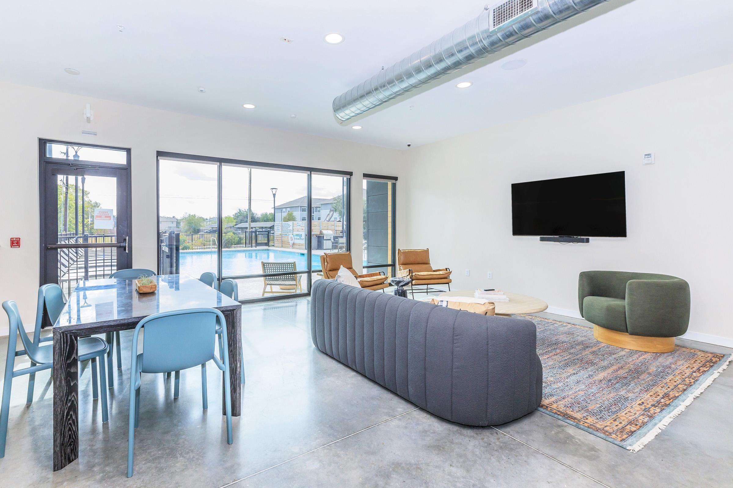A modern living space featuring a gray sectional sofa, a round wooden coffee table, and two armchairs. A dining table with blue chairs is visible. Large windows showcase a pool area outside. The walls and floor have a neutral color palette, creating a bright and inviting atmosphere.