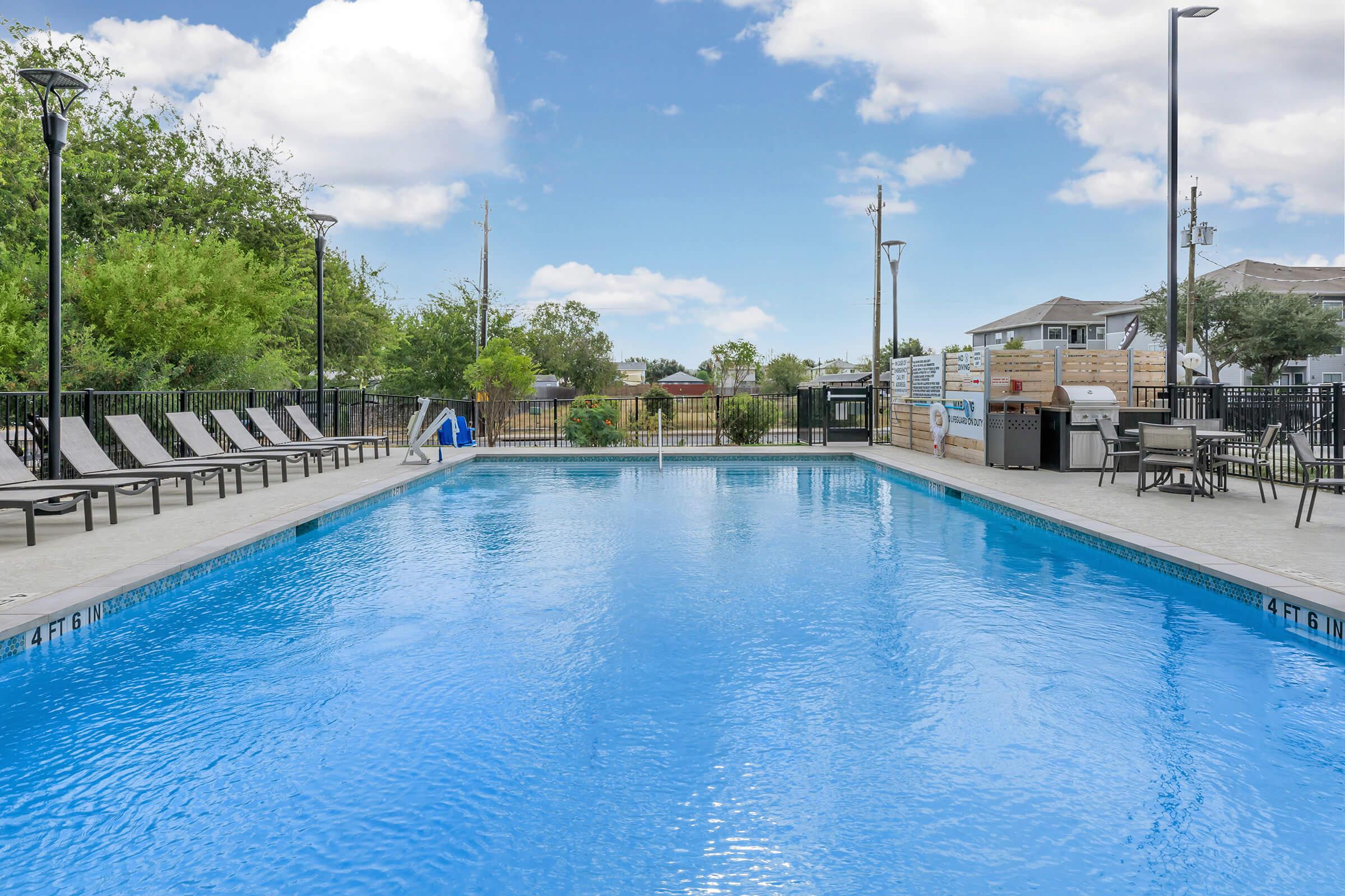 A clear blue swimming pool surrounded by lounge chairs, with trees and buildings in the background. The pool area is well-lit with lampposts, and a poolside grill is visible. The inviting scene suggests a relaxing outdoor space for leisure or social gatherings.