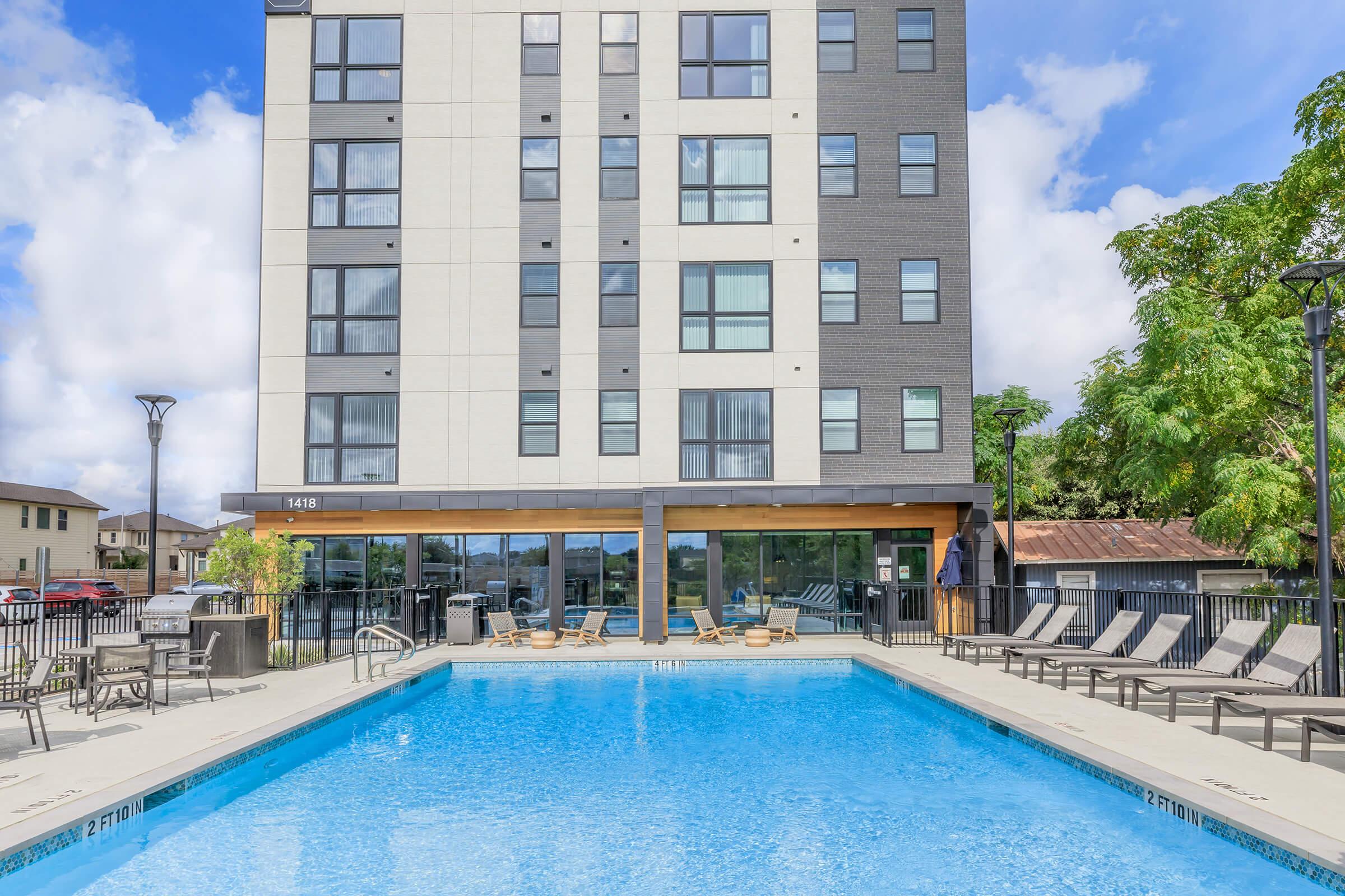 A modern apartment building with a large outdoor swimming pool in front. The pool area features lounge chairs, surrounded by a fence and landscaping. The building has multiple floors with large windows, and the sky shows a few fluffy clouds.