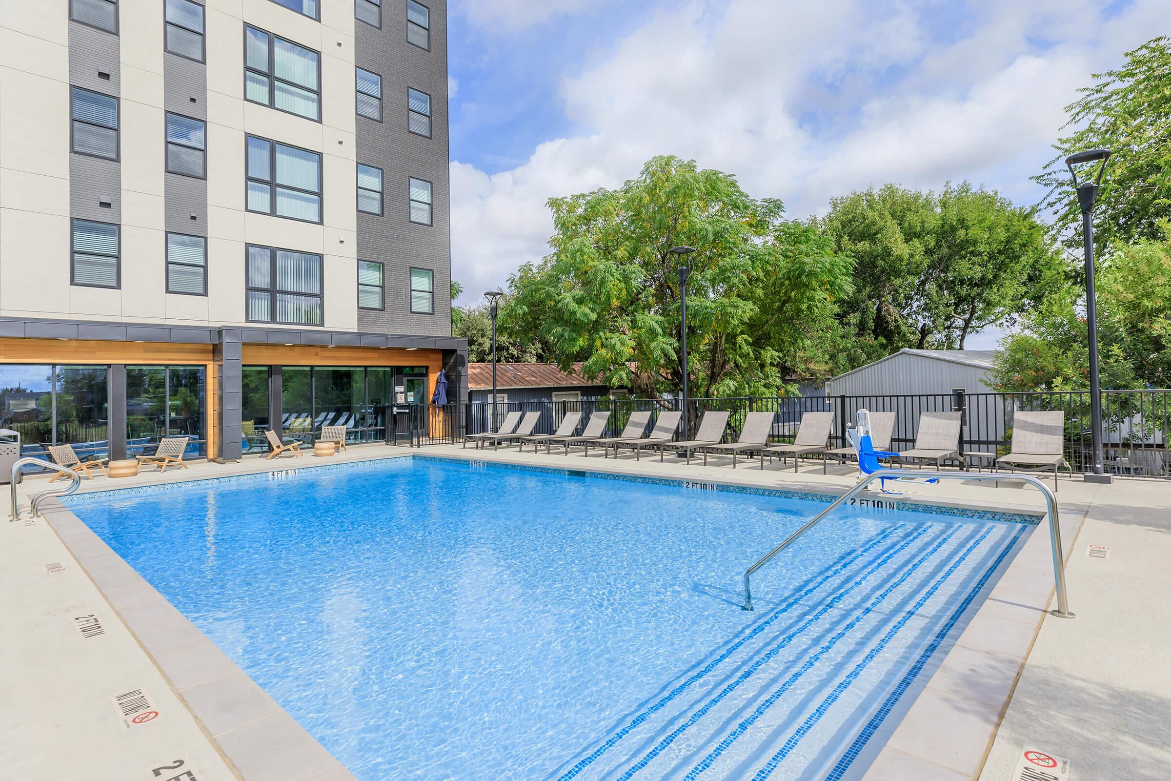 A clear swimming pool surrounded by lounge chairs, located in the courtyard of a modern building. Trees and greenery are visible in the background, under a partly cloudy sky. The pool area features a ladder for easy access, and the building shows multiple windows and a contemporary design.