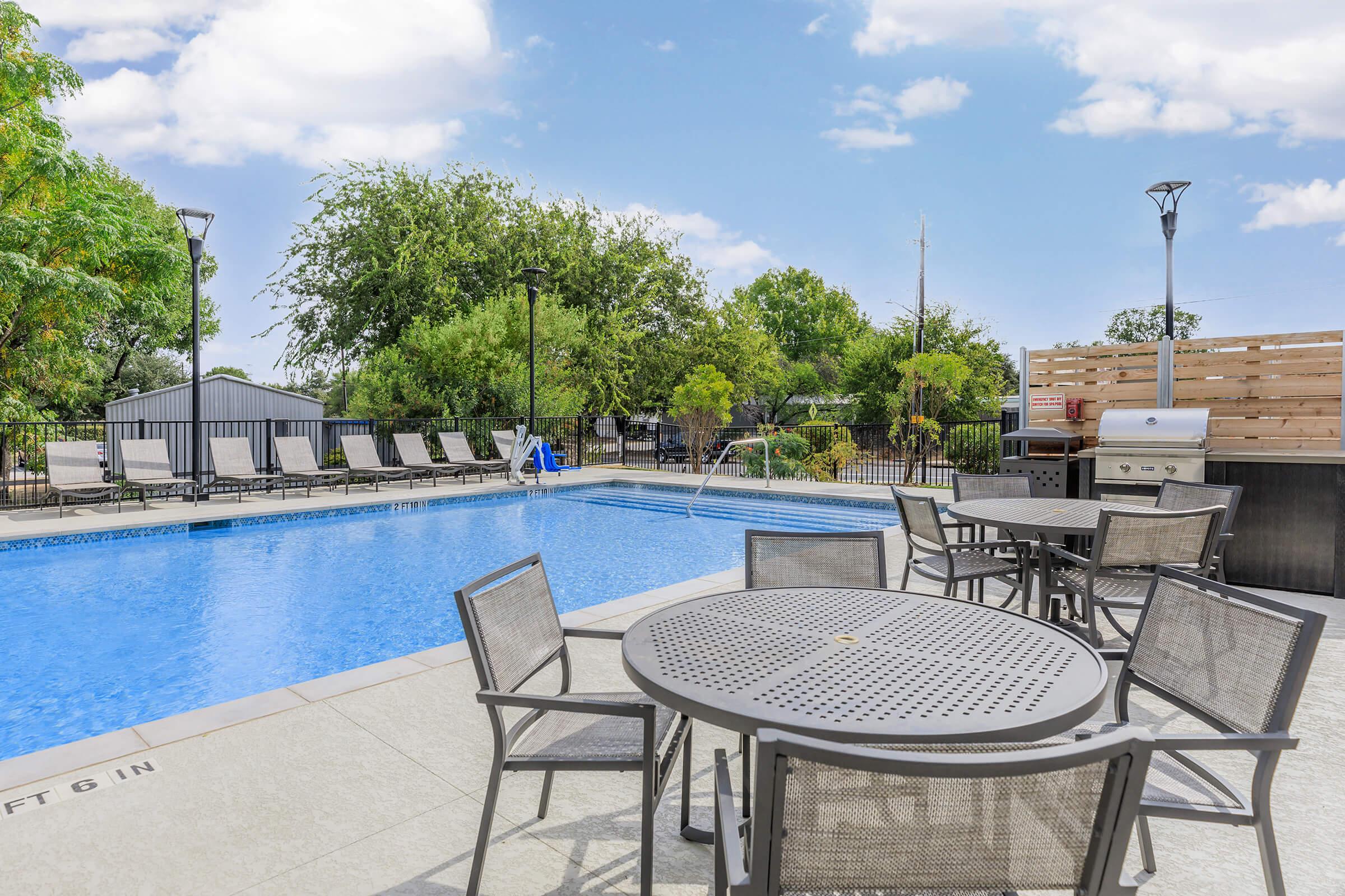 A clear swimming pool surrounded by patio furniture, including metal tables and chairs. Lush green trees and shrubs are visible in the background, along with a barbecue grill nearby. The scene is set under a bright sky with a few clouds, creating a welcoming outdoor atmosphere.