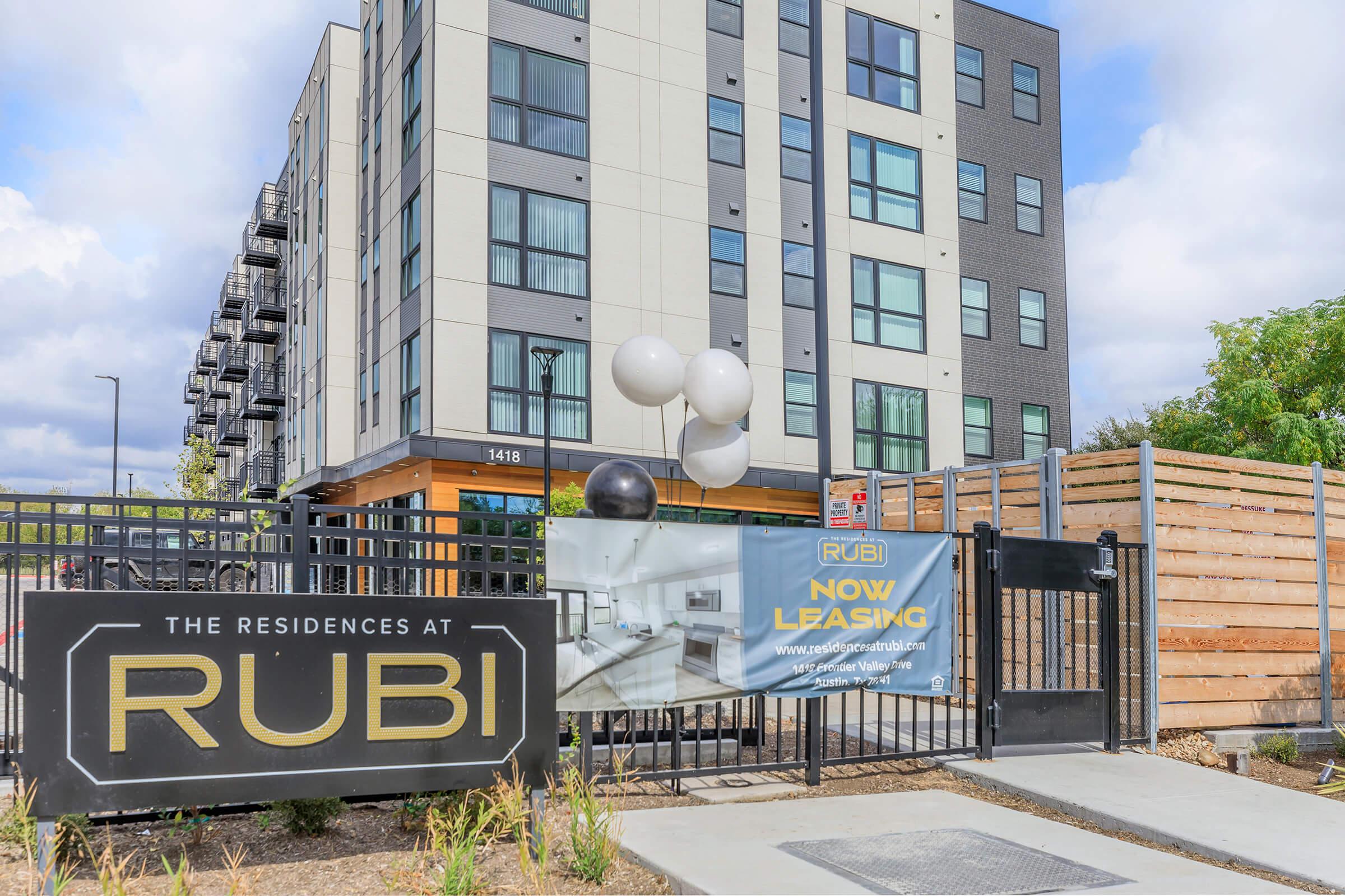 A modern apartment building with a sign that reads "The Residences at RUBI" and a banner stating "NOW LEASING." The entrance features a gated area with decorative balloons and landscaping. The building has multiple stories and a contemporary design, with windows and balconies visible.