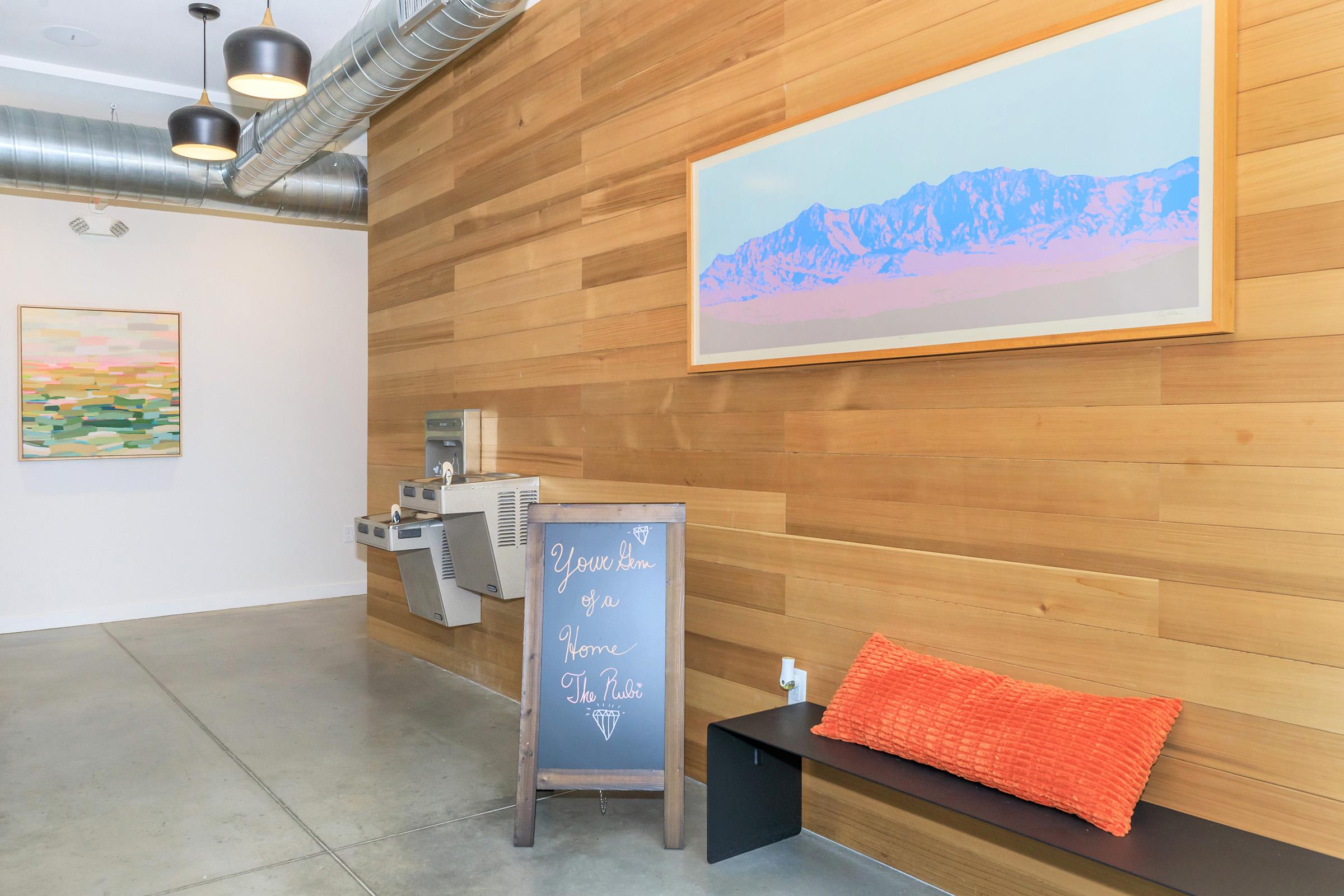 Interior view of a modern space featuring a wooden wall, a wall-mounted water fountain, and a stylish art piece depicting mountains. A chalkboard sign reads "Welcome as a Home to the Public," and there's a bench with an orange cushion. The floor is concrete, and the lighting is industrial.