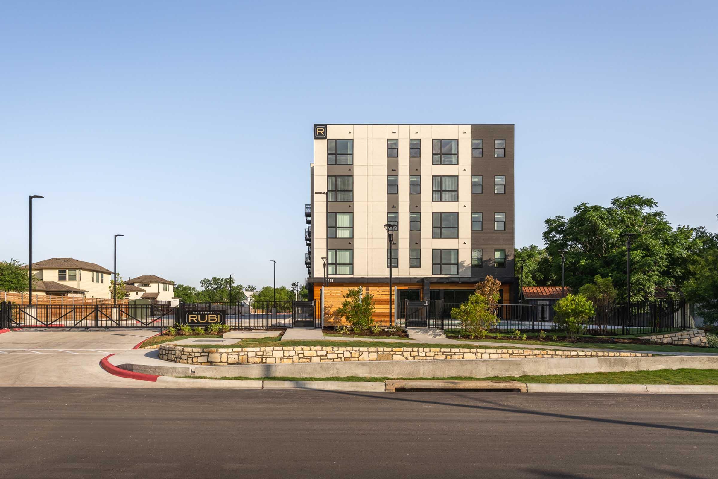 A modern multi-story building with a sleek design and a welcoming entrance. The façade features a combination of light and dark materials. In the foreground, there is a landscaped area with trees and shrubs, and the building is surrounded by a gated perimeter. Clear blue sky in the background.