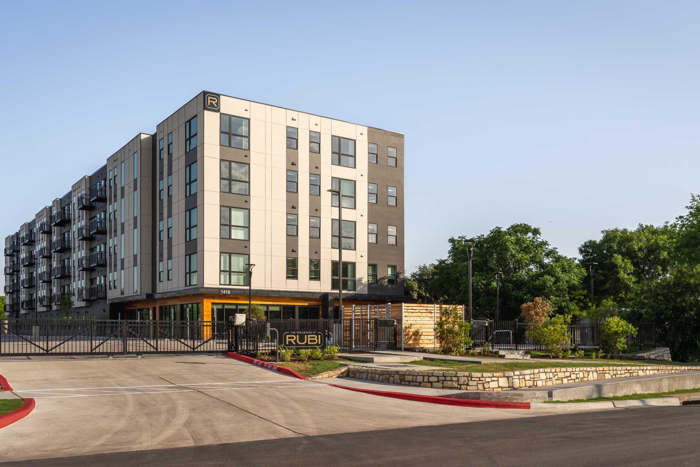 Modern multi-story apartment building with a sleek design featuring large windows and balconies. The entrance is gated, and there are landscaped areas with trees and bushes nearby. The building has a prominent sign displaying the name "RUBI." The scene is set in a clear, sunny environment.