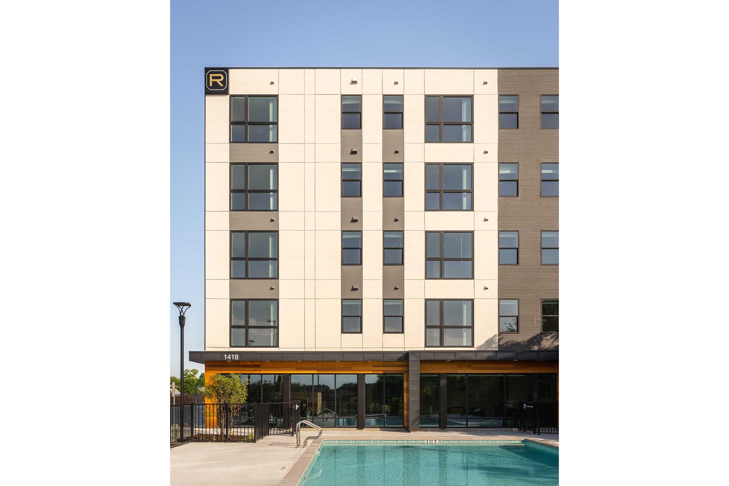 Modern building exterior with large windows and a pool in the foreground. The structure features a mix of light and dark materials, with a prominent sign displaying an "R". The facade is sleek and contemporary, set against a clear blue sky. The pool area includes a deck and a ladder for access.