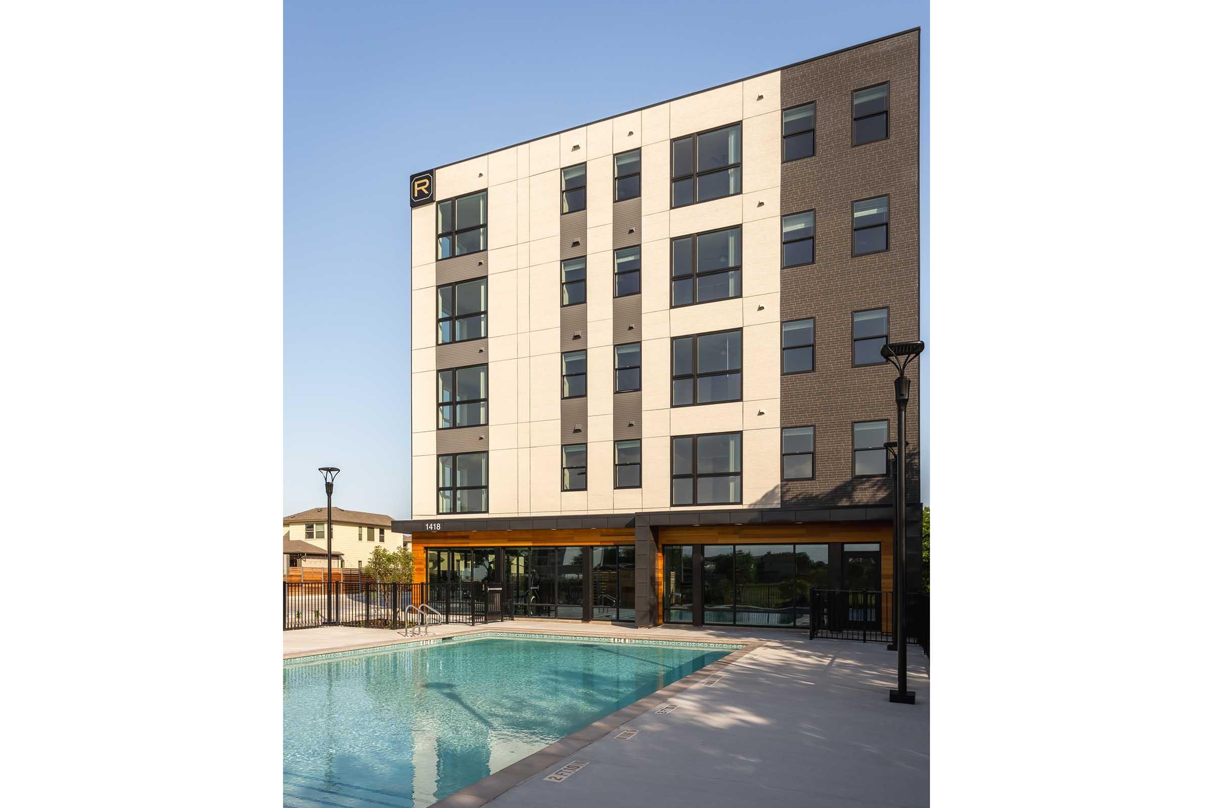 Modern multi-story building with large windows, featuring a ground-level pool area. The facade combines light and dark materials, and there are outdoor light fixtures surrounding the pool. The building is located in an urban setting, with a clear blue sky overhead.