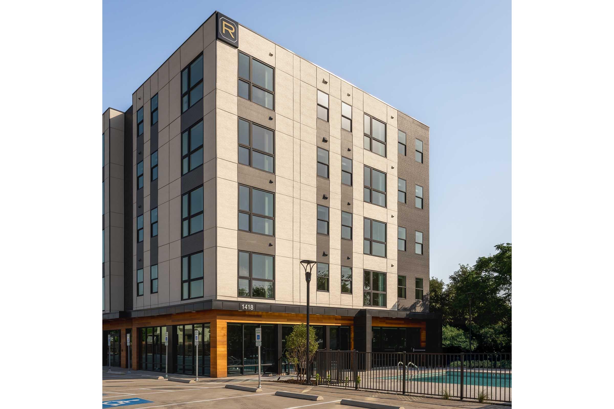 Modern multi-story building with a clean architectural design featuring large windows. The exterior combines light and dark materials, and there is a canopy at the entrance. A fenced pool area is visible, along with parking spaces in front. The scene is set against a clear blue sky.