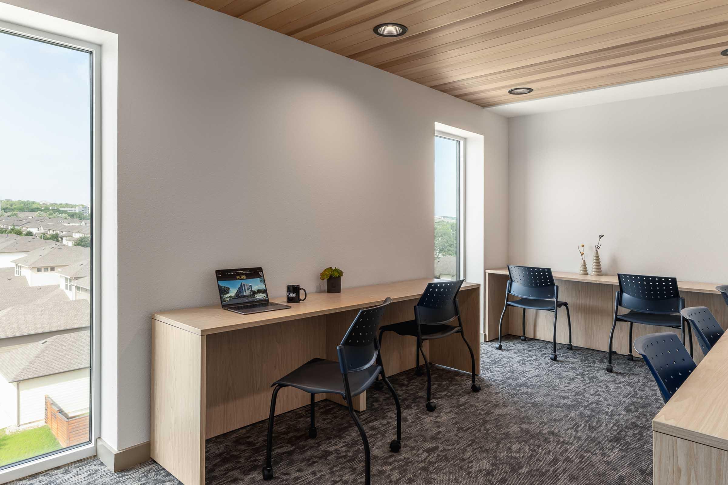 A modern study or work space featuring a light wood desk against a wall with two windows. Each desk has a chair, and one desk holds a laptop with a small potted plant. The decor is minimalist, with a neutral color palette and a view of rooftops outside the windows.