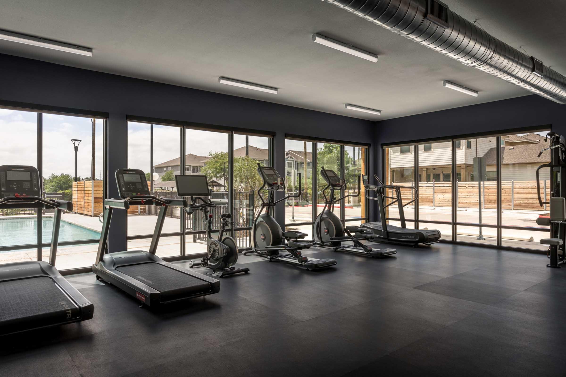 A modern gym interior featuring multiple cardio machines, including treadmills and ellipticals, arranged neatly on a black floor. Large windows allow natural light to illuminate the space, offering views of an outdoor pool area. The walls are painted in dark tones, creating a stylish and focused workout atmosphere.