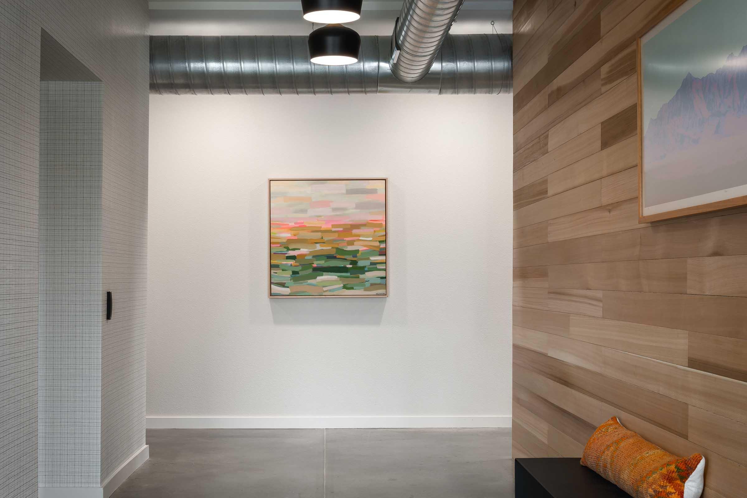 A modern interior hallway featuring a wooden accent wall and a framed abstract painting in soft pastel colors. The space has gray concrete flooring and industrial-style lighting. A small decorative pillow is placed on a dark bench, enhancing the contemporary aesthetic.