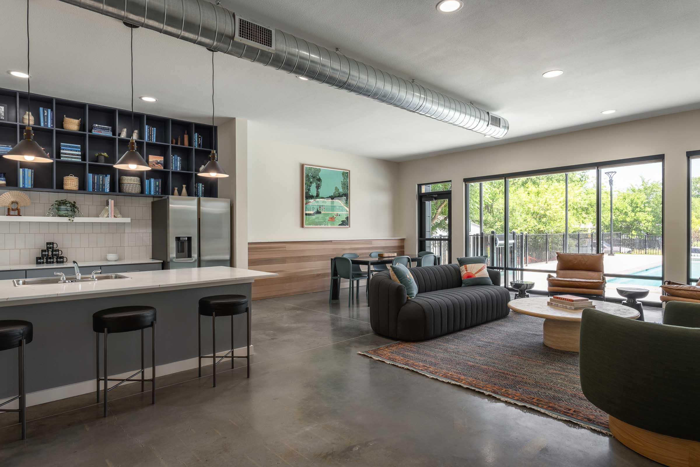 A modern indoor lounge area featuring a kitchen island, bar stools, and a comfortable seating arrangement with sofas and chairs. Large windows offer views of an outdoor pool area, and the decor includes bookshelves and artwork, creating a stylish and inviting atmosphere.
