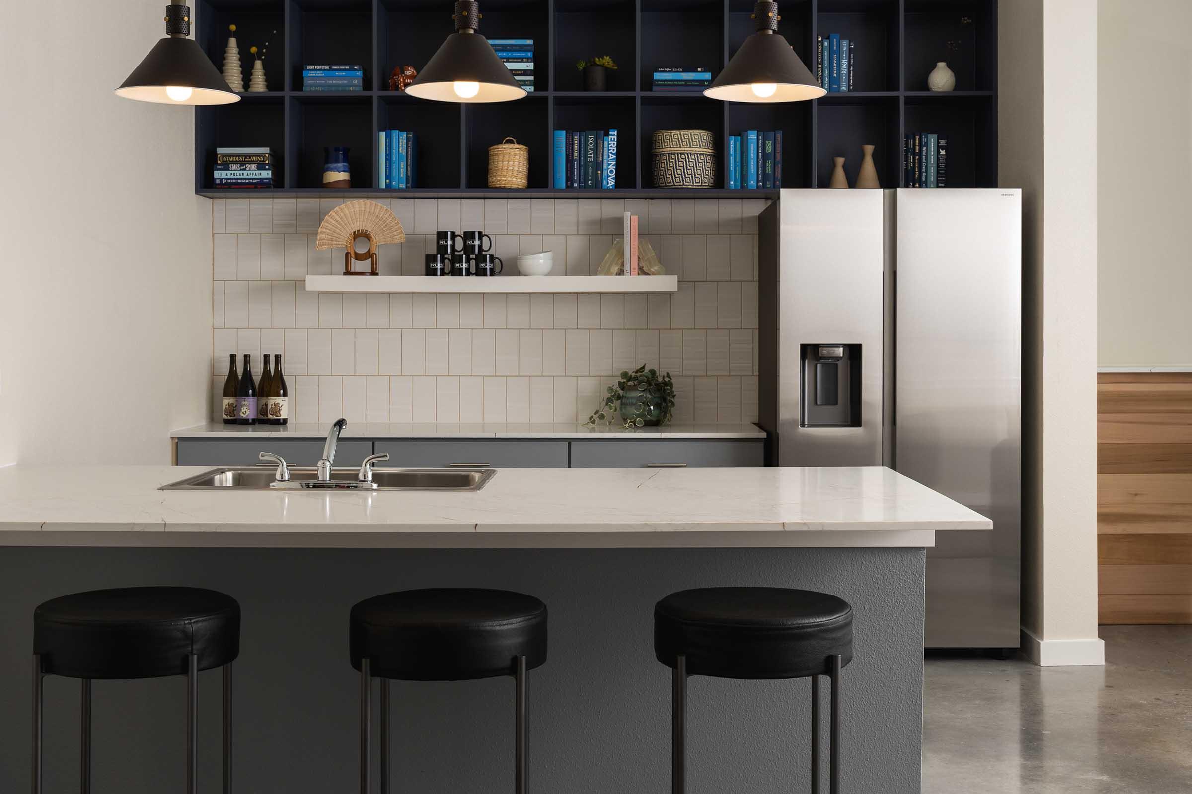 A modern kitchen featuring a large island with a white marble countertop and three black stools. The background includes dark shelving with books and decorative items, and a stainless steel refrigerator. Pendant lights hang above the island, adding contemporary flair to the space.