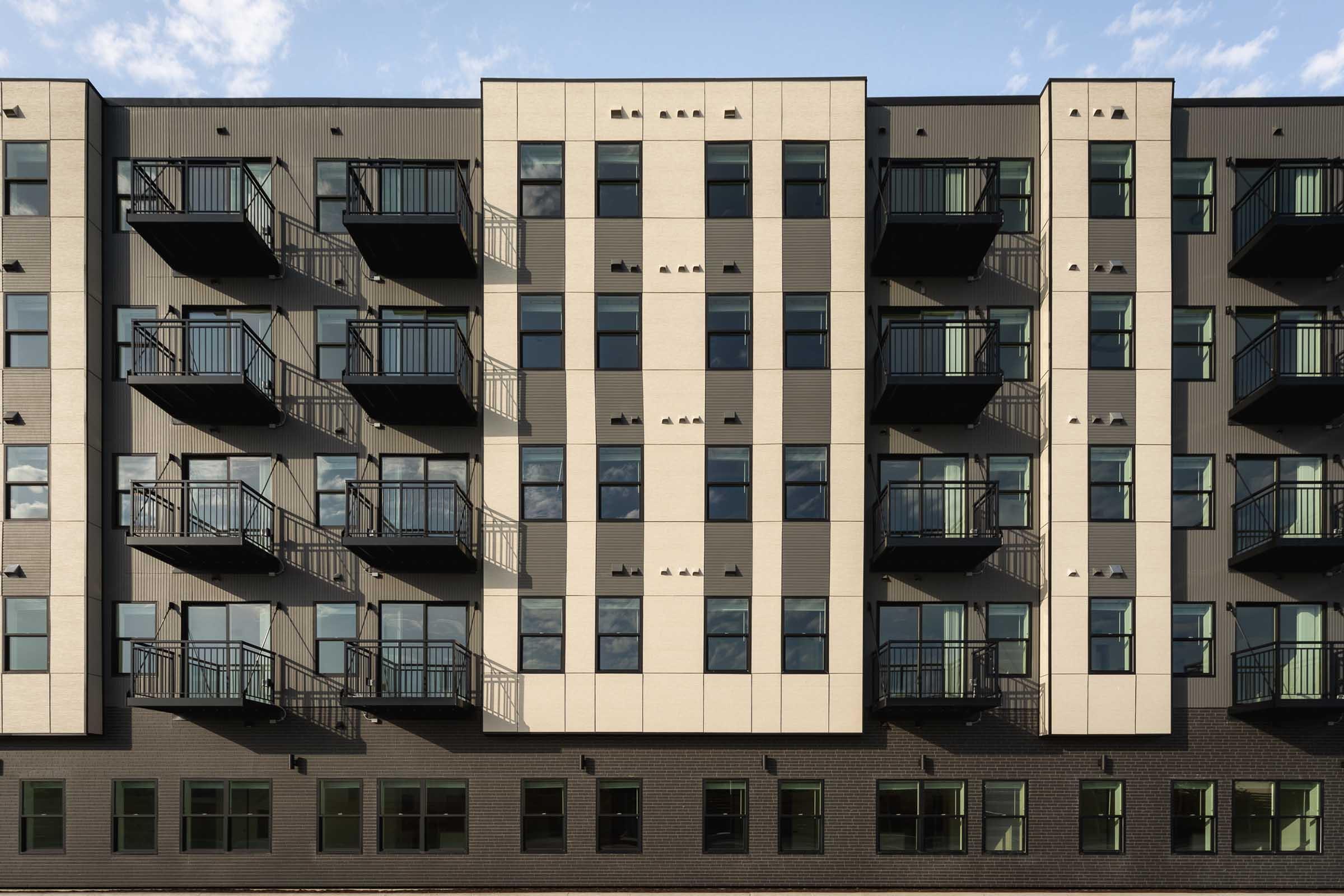 A modern apartment building facade featuring a combination of light and dark-colored panels. The structure has multiple balconies with metal railings and large windows. The design includes a symmetrical arrangement with sections that highlight vertical and horizontal elements, set against a blue sky.