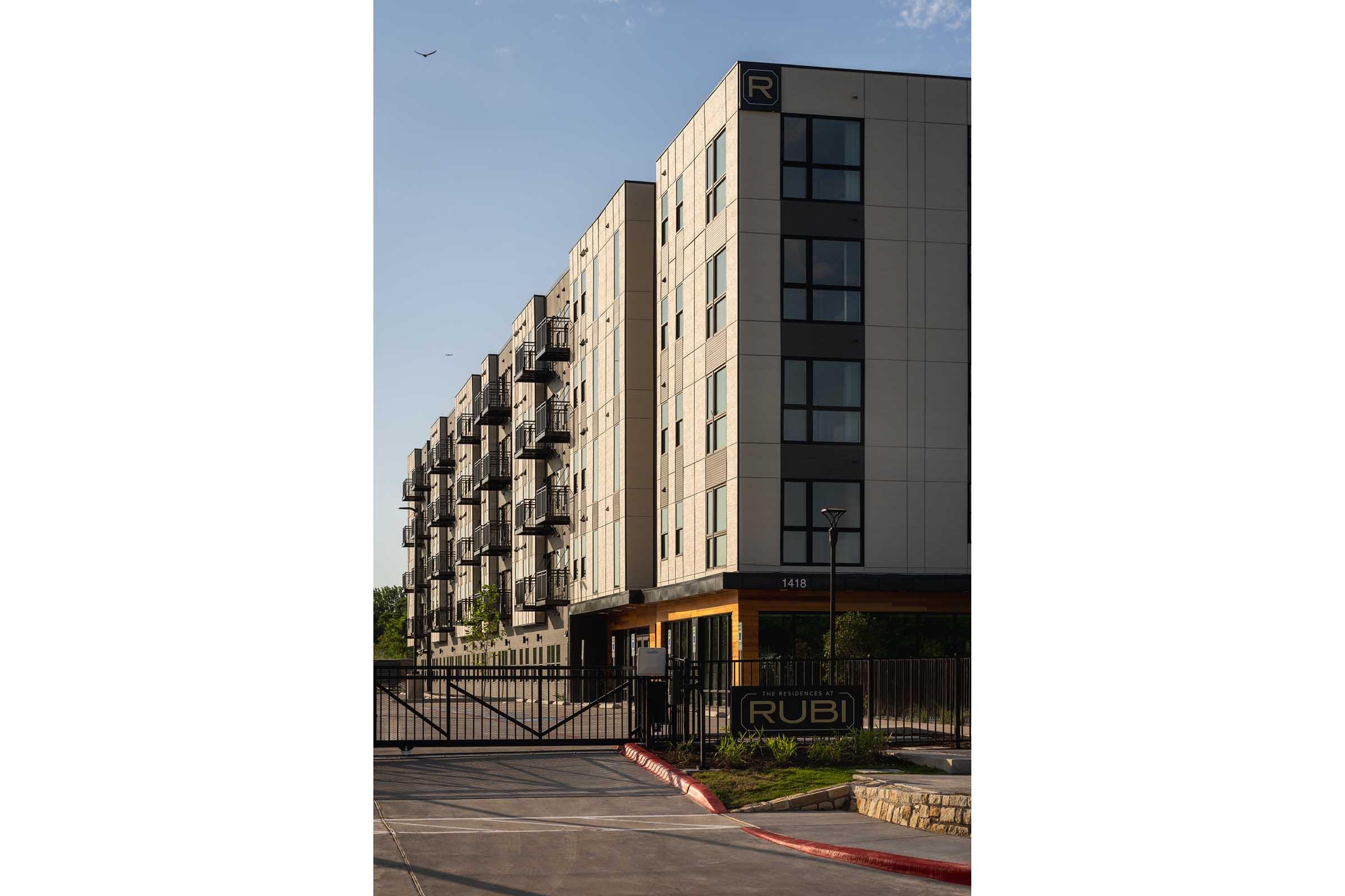Modern apartment building with a sleek design, featuring multiple floors, balconies, and a gated entrance. The building is labeled "RUBI" and is set against a clear sky with a hint of greenery in the surroundings.