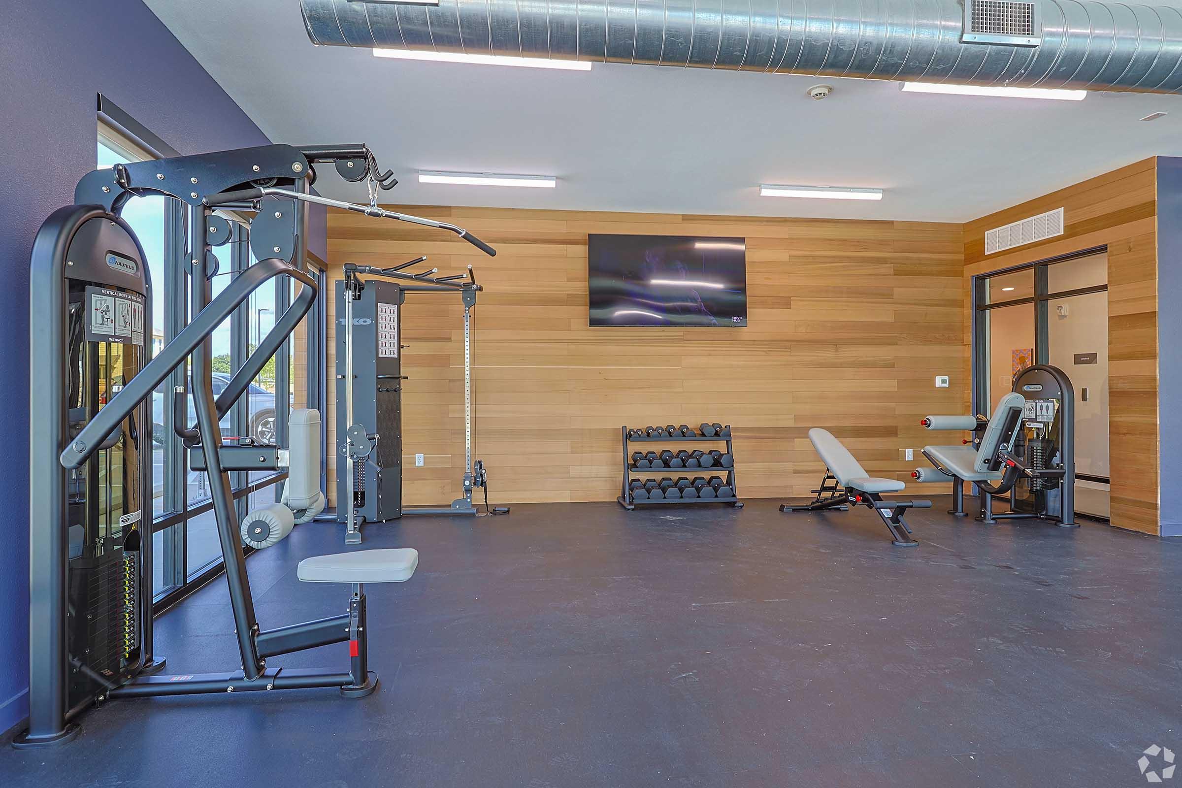 A modern gym interior featuring workout equipment, including a cable machine, a weight bench, and a rack of dumbbells. The walls are finished in wood, and there is a flat-screen TV mounted on the wall. Large windows provide natural light to the space.