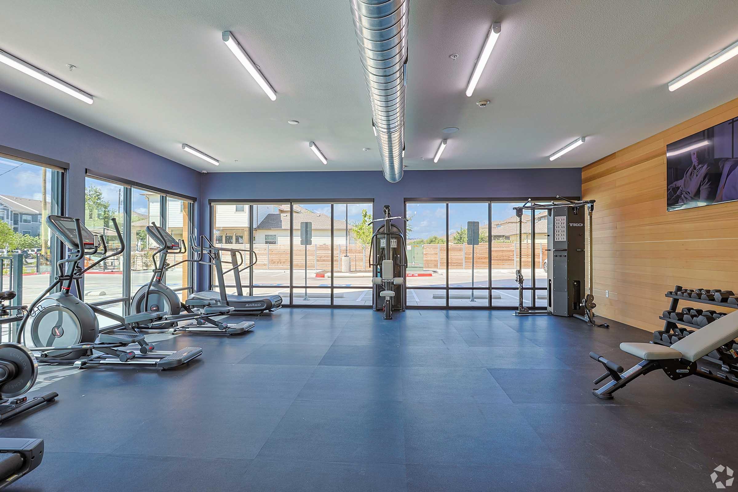 A modern gym interior featuring various exercise equipment, including ellipticals and strength training machines, with large windows allowing natural light. The space has a clean, minimalist design with wooden accents and an outdoor view.