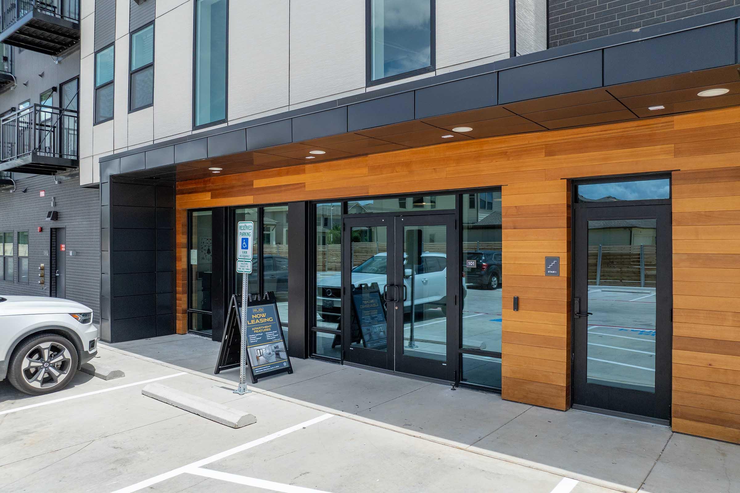 Modern building facade featuring a wooden accent wall and large glass doors. A menu board is displayed outside, and there are parking spaces visible in front. The building has a contemporary design with a mix of materials, including black panels and brick. A vehicle is parked nearby.