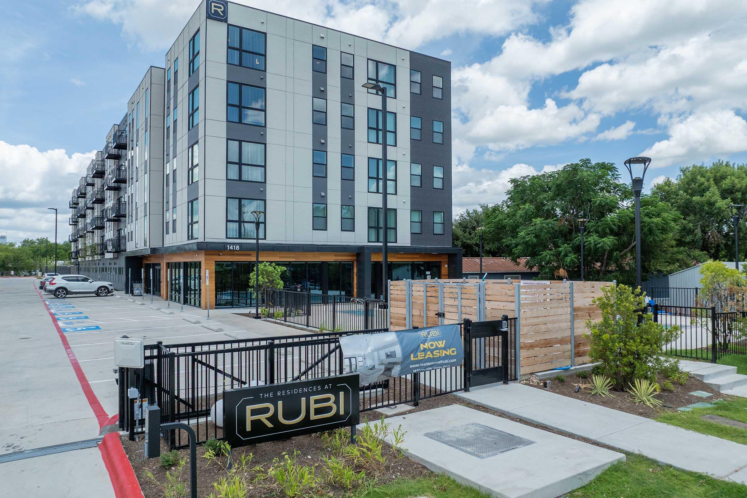 A modern apartment building with a five-story design featuring a mix of dark and light exterior panels. In front, there is a gated entrance with a leasing sign. The parking lot is visible with several cars parked, and there are trees and green landscaping around the property under a partly cloudy sky.