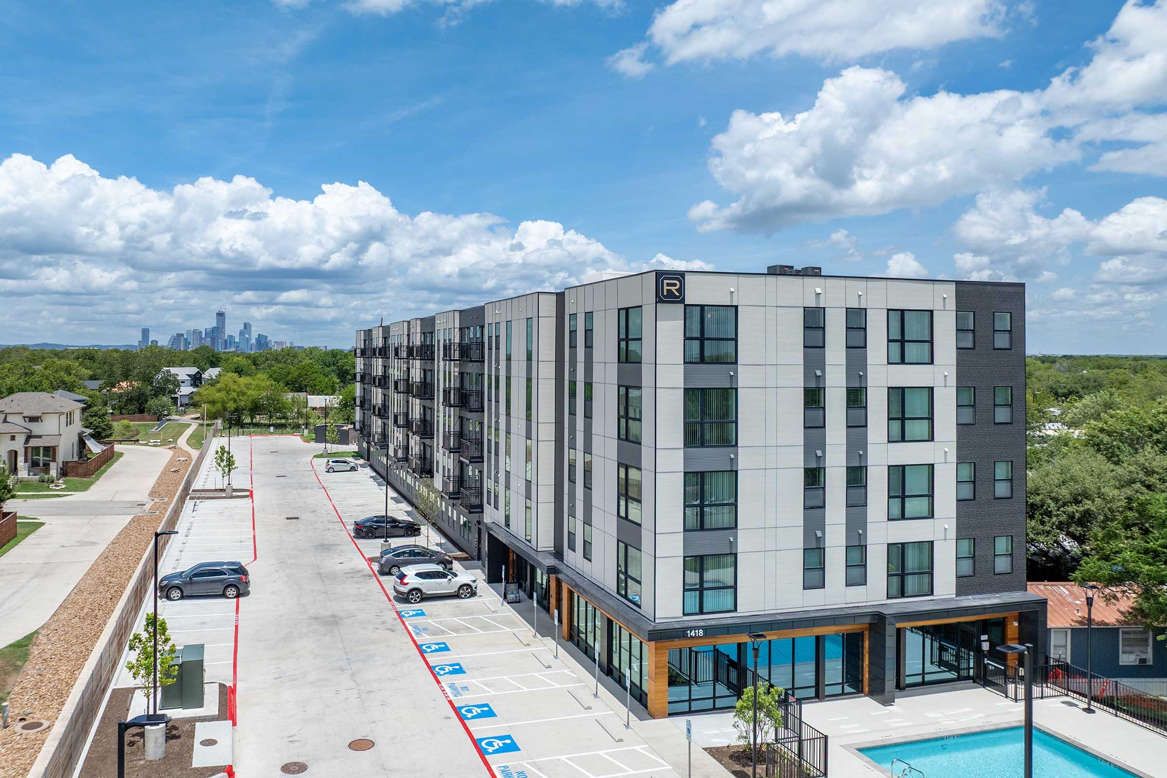 A modern multi-story apartment building with large windows and a pool area, surrounded by green trees and a parking lot. In the background, a city skyline is visible under a partly cloudy sky. The setting conveys a contemporary urban living environment.