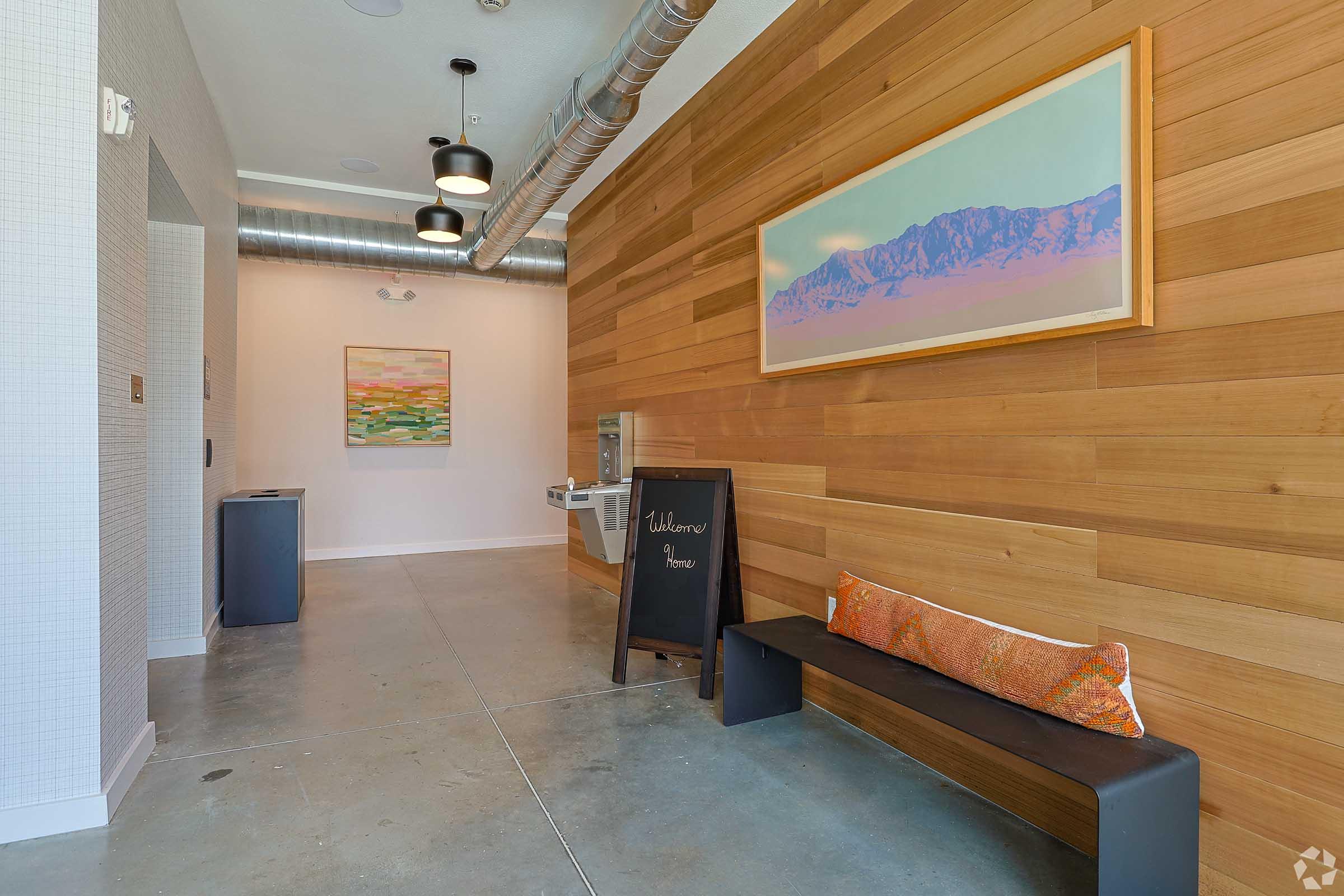 A modern and stylish entrance area featuring a wooden accent wall, a decorative painting, a small bench with a colorful cushion, a chalkboard sign welcoming visitors, a water cooler, and a clean concrete floor. The space has a bright and inviting atmosphere, enhanced by natural light.