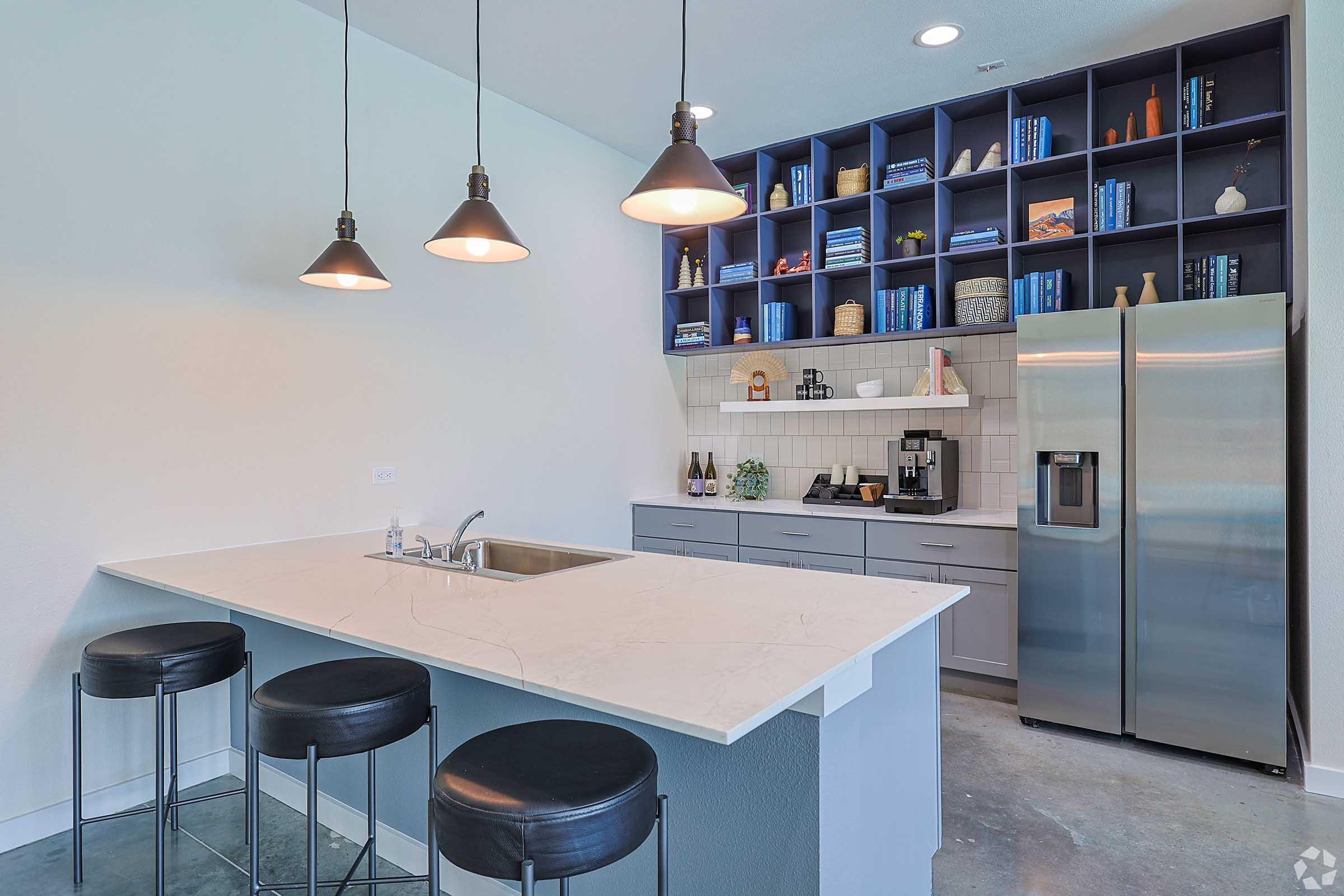 Modern kitchen featuring a large island with a sink and three black bar stools. Above the island hang three pendant lights. In the background, there are shelves filled with books, decorative items, and a coffee maker, alongside a stainless steel refrigerator and grey cabinetry. The floor is polished concrete.