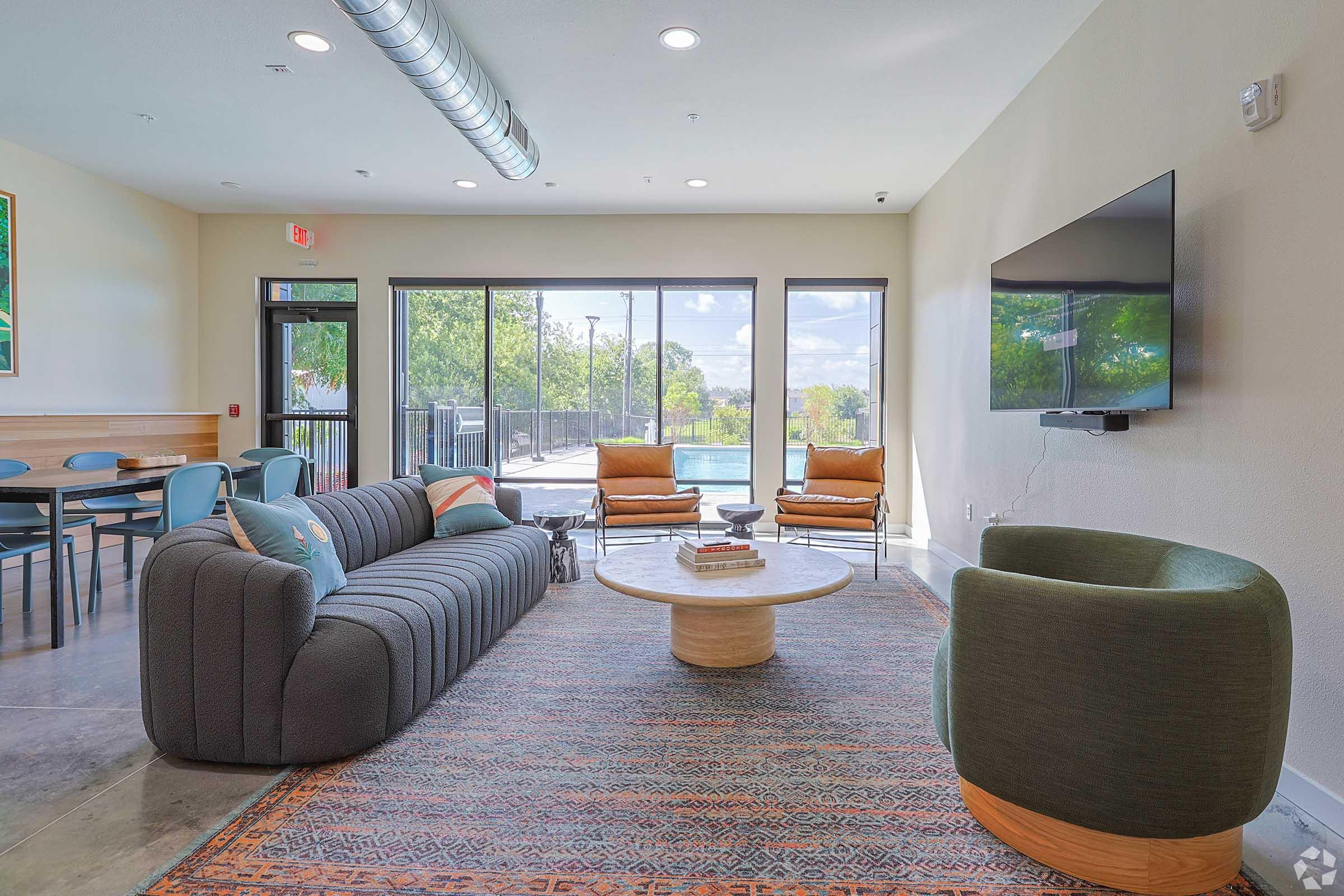 A modern interior space featuring a cozy gray sofa, a round wooden coffee table, and two green armchairs. Large windows reveal a view of a pool area outside. The room has a stylish rug and is well-lit with natural light, creating a welcoming atmosphere.