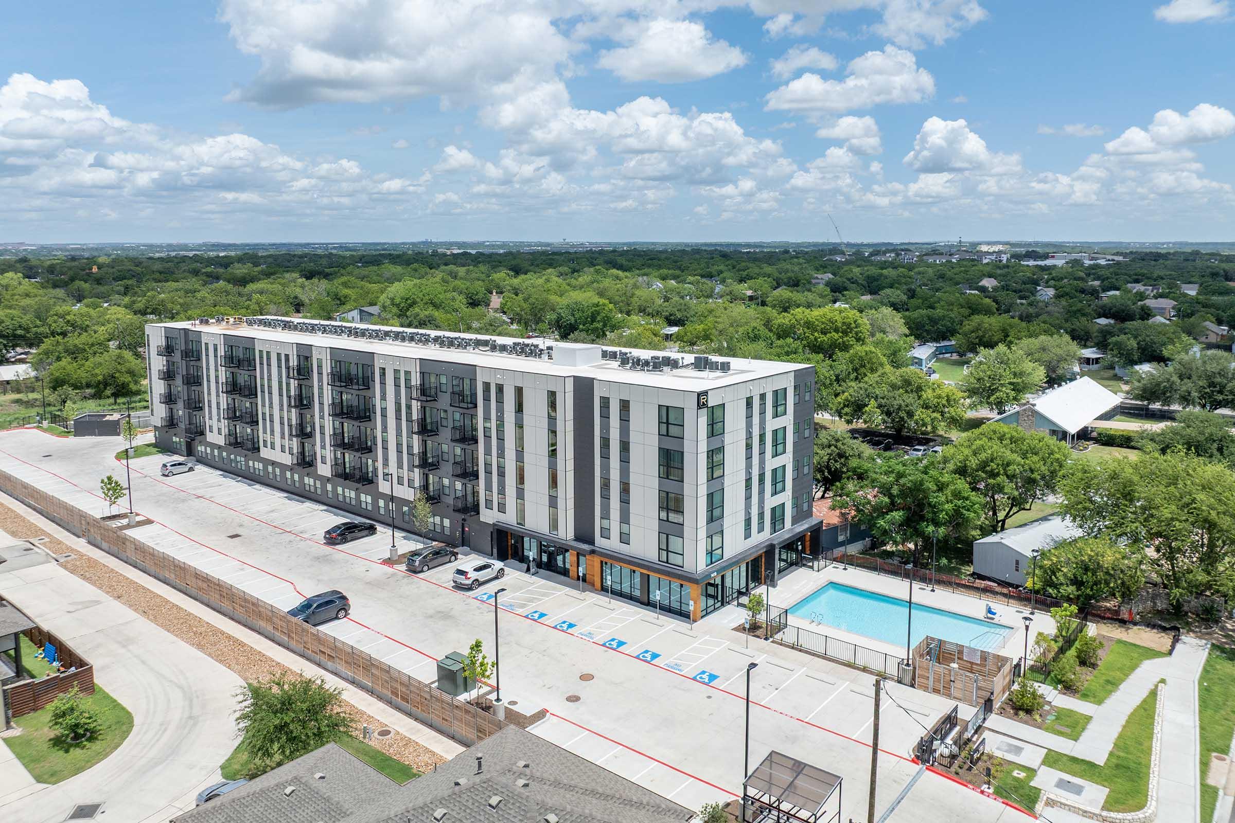 A modern multi-story residential building with balconies, situated in an urban area surrounded by greenery. In front, there’s a parking lot with a few cars and a swimming pool area. The sky is partly cloudy, and the view reveals a mix of foliage and buildings in the distance.