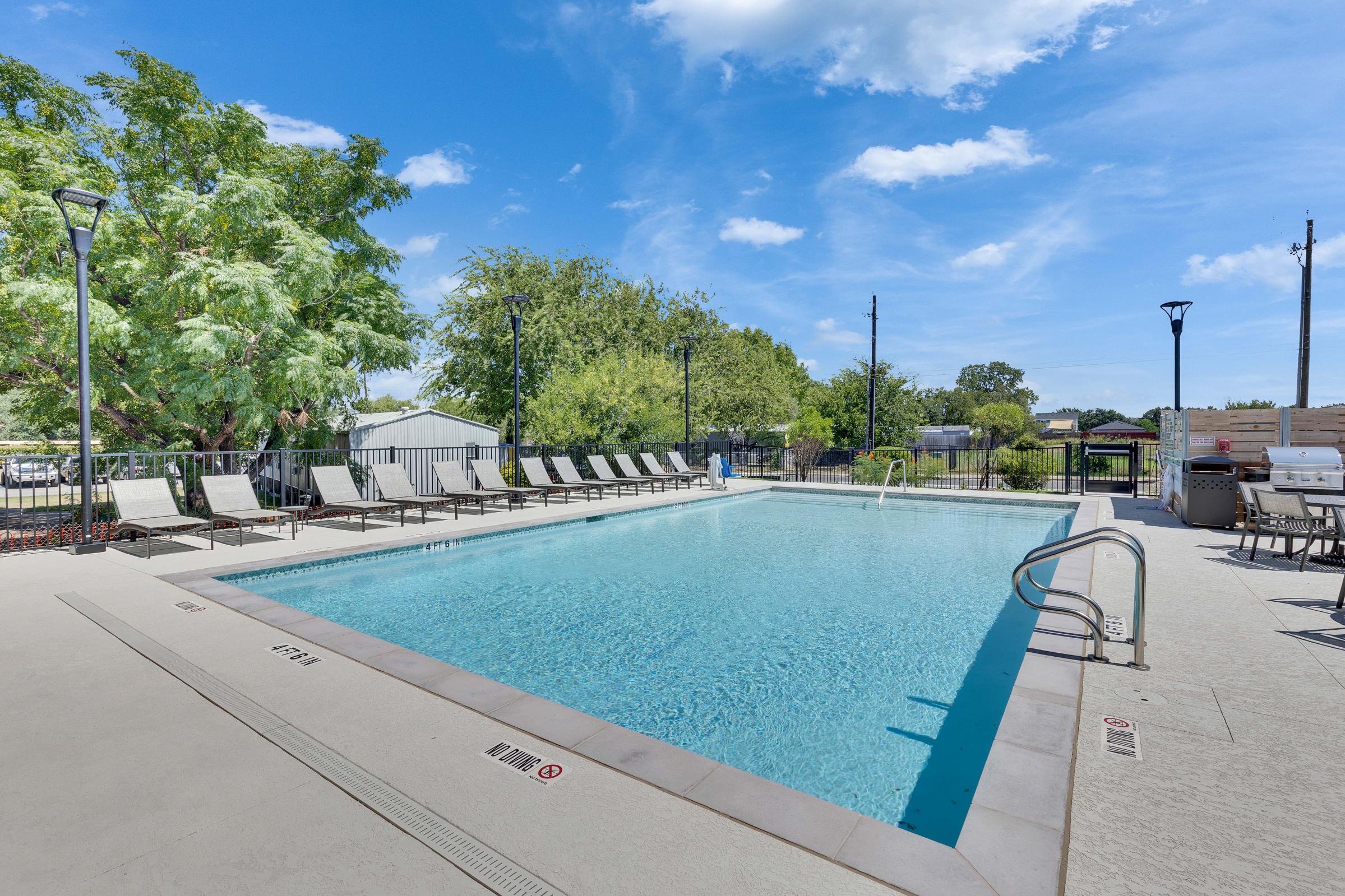 A clear blue swimming pool surrounded by empty lounge chairs, with green trees in the background and a bright blue sky. The pool area features a smooth, light-colored deck and several lamp posts, creating a welcoming outdoor space for relaxation and leisure.