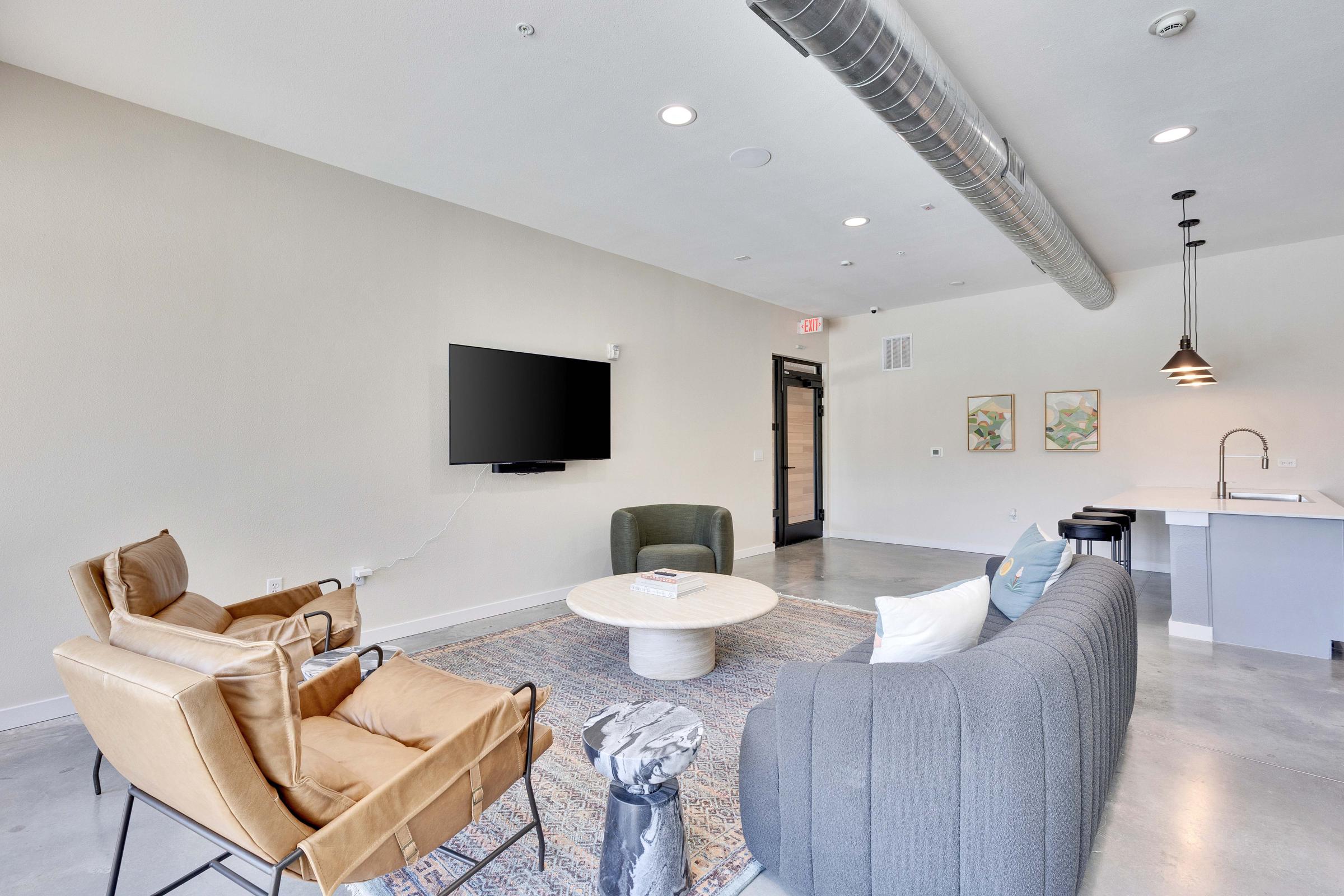A modern living space featuring two stylish armchairs, a gray sofa, and a circular coffee table on a patterned rug. A wall-mounted television is visible opposite the seating area, with a kitchen bar in the background. The room has a bright, open feel with minimalistic décor and neutral colors.