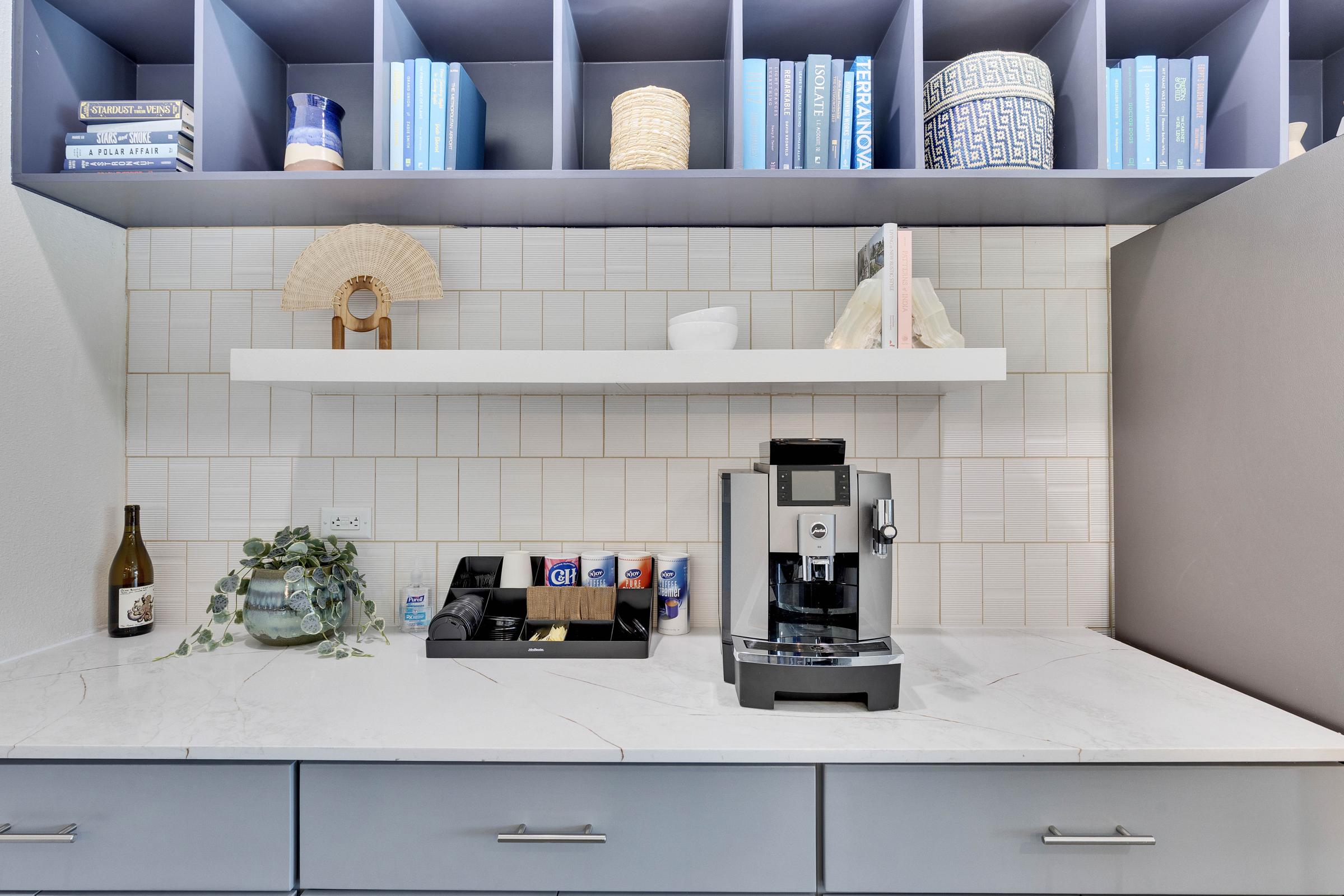 A modern kitchen counter featuring a coffee machine, a selection of beverages, a decorative plant, and a wine bottle. Above, there's a shelf holding books and a decorative vase. The wall is tiled with soft-colored tiles, and there is a stylish light fixture providing ambient lighting.