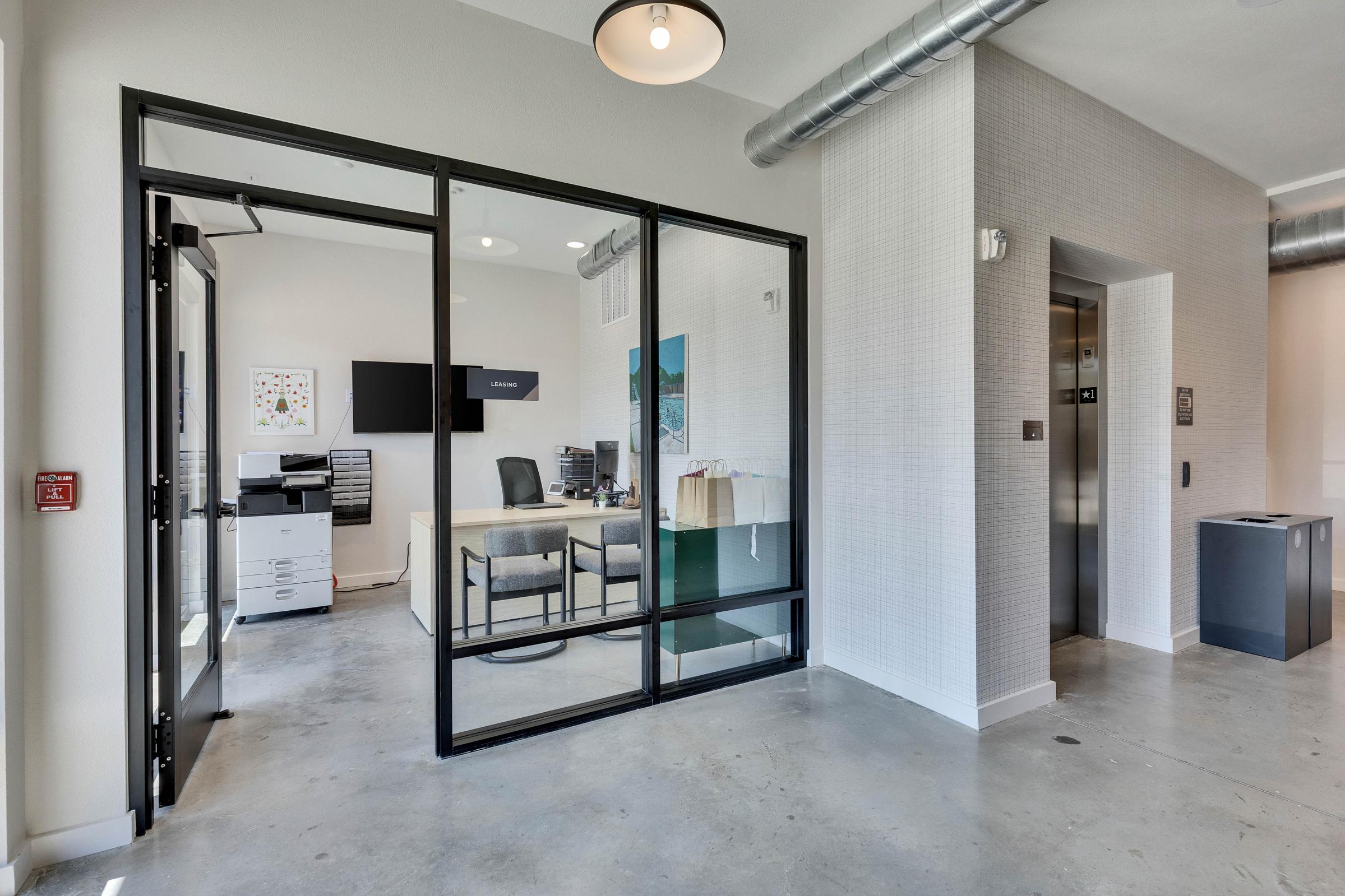 A modern office space featuring glass walls, a desk with a computer, and an office chair. There’s a printer nearby and an elevator in the background. The floor is concrete, and the space has contemporary lighting, creating a bright and professional atmosphere.