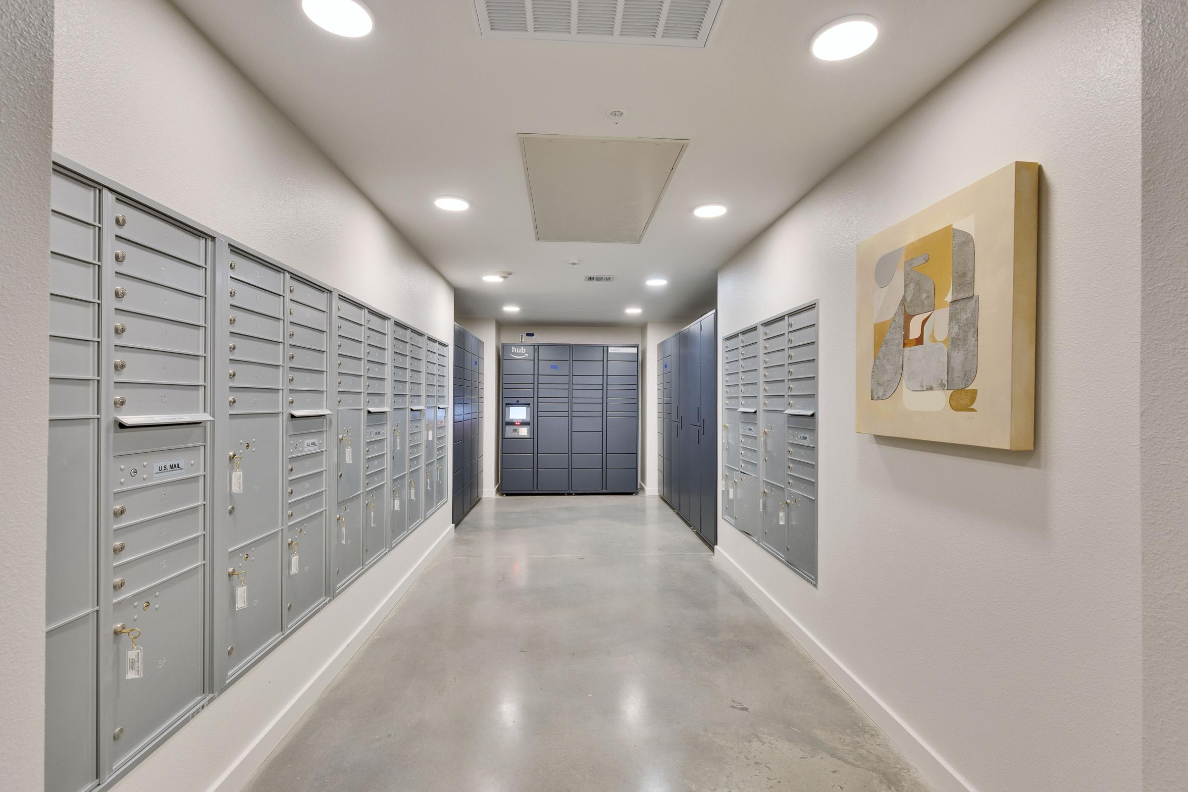 A narrow corridor featuring gray mailbox units lined against the walls, with a larger package locker unit at the end. The floor is polished concrete, and a modern piece of artwork hangs on the right wall, adding a touch of color to the otherwise neutral space. Bright overhead lights illuminate the area.