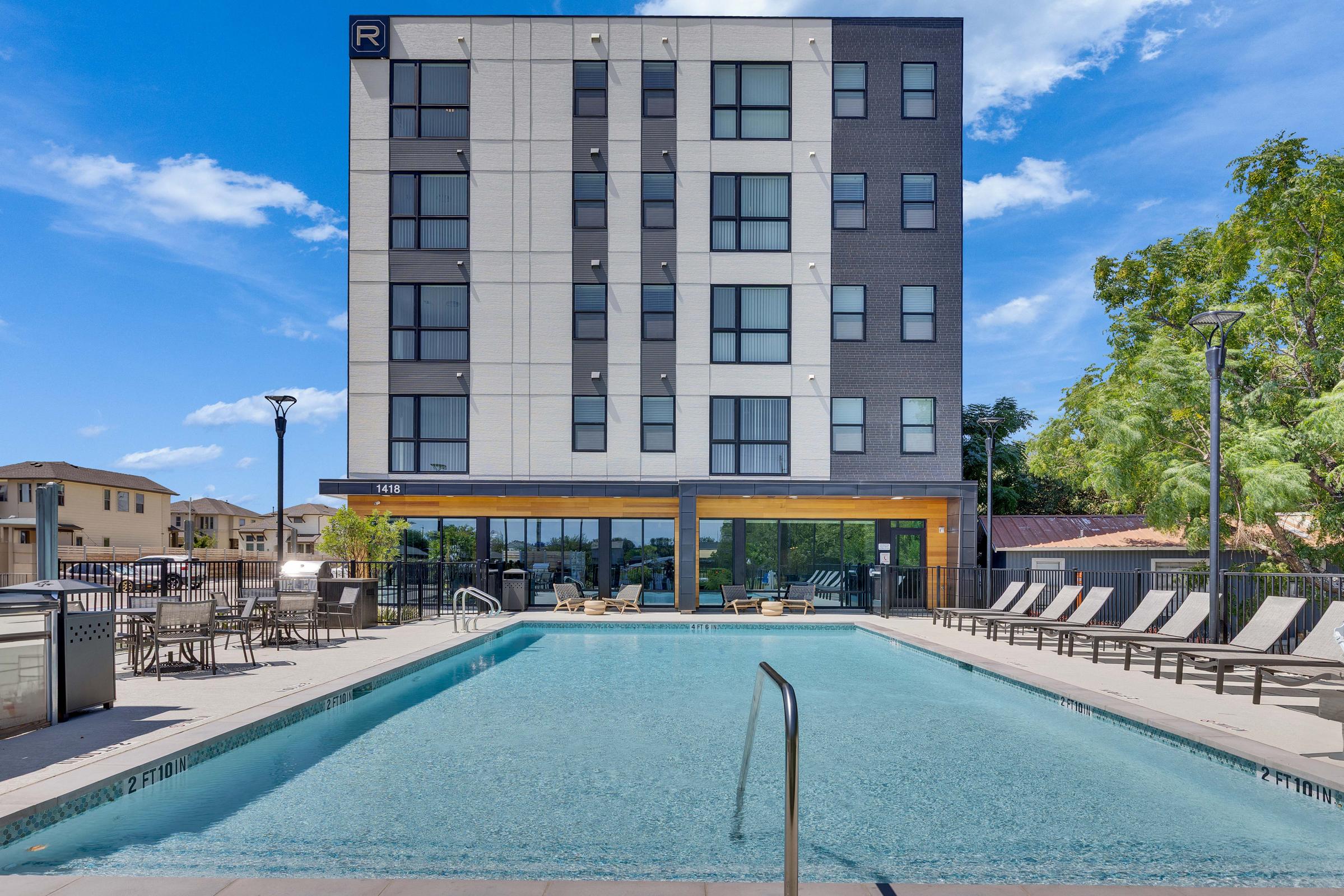 A modern hotel building with a sleek design, featuring large windows and a prominent entrance. In the foreground, there is a swimming pool surrounded by lounge chairs and a patio area. The sky is clear with a few clouds, and there are trees and other buildings in the background, creating a welcoming outdoor space.