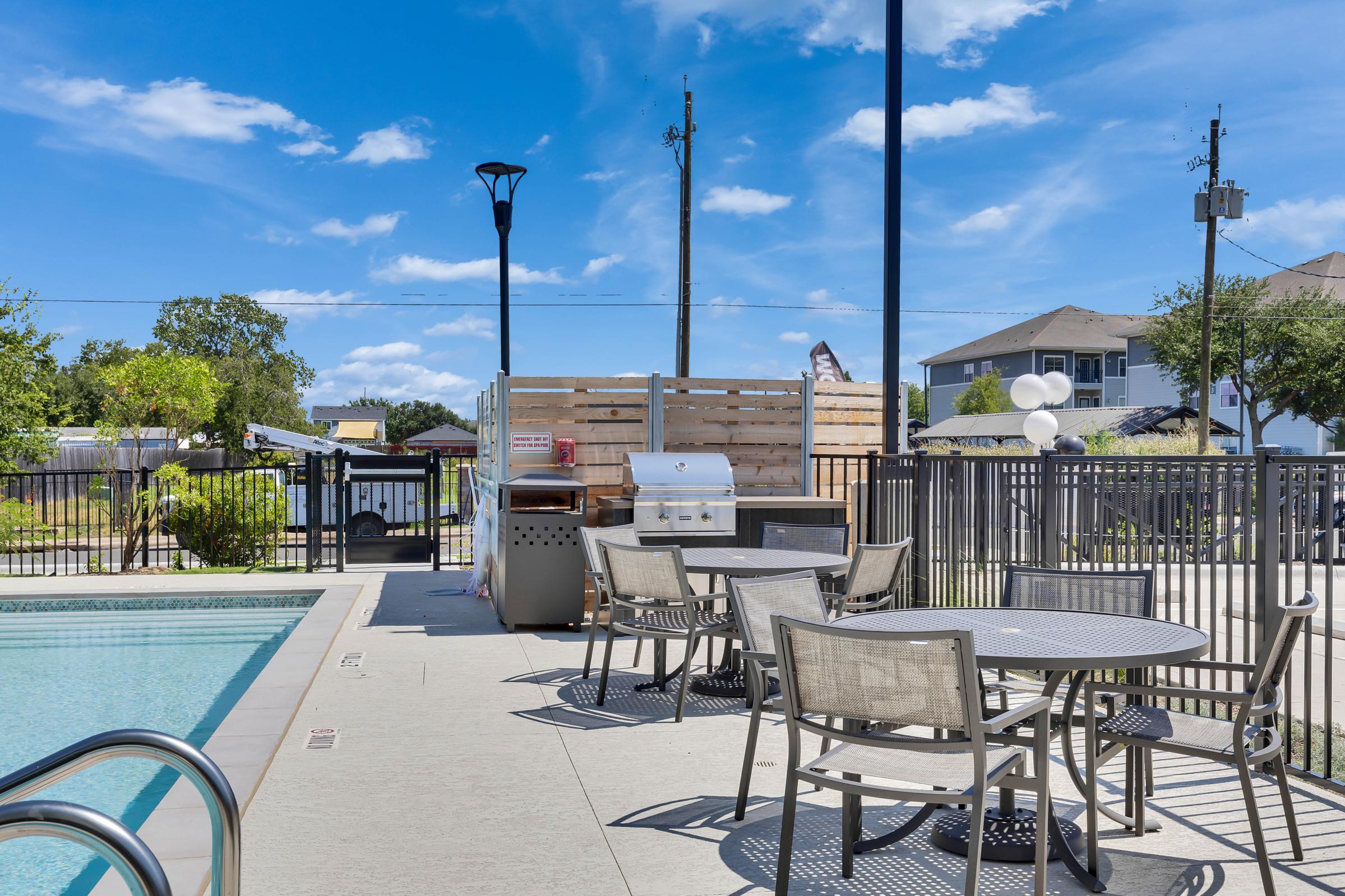 A clear blue sky with a few clouds above a well-maintained outdoor pool area. The scene features a poolside grill station, outdoor tables and chairs, and a landscaped area with trees. Nearby, a fenced-in yard and apartment buildings can be seen in the background.
