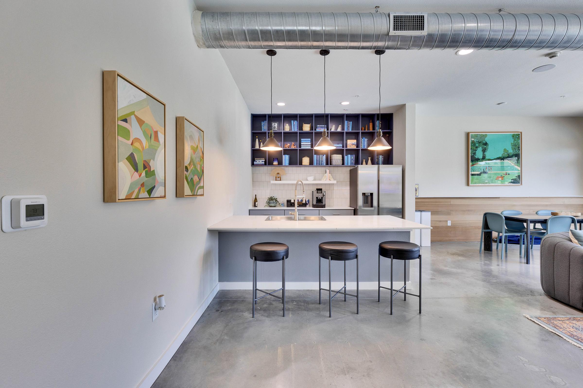 Modern kitchen with a minimalist design featuring a white countertop and black bar stools. There are two framed art pieces on the wall, a blue bookshelf filled with books, and pendant lighting above the counter. A stainless steel refrigerator is visible, along with a dining area in the background.