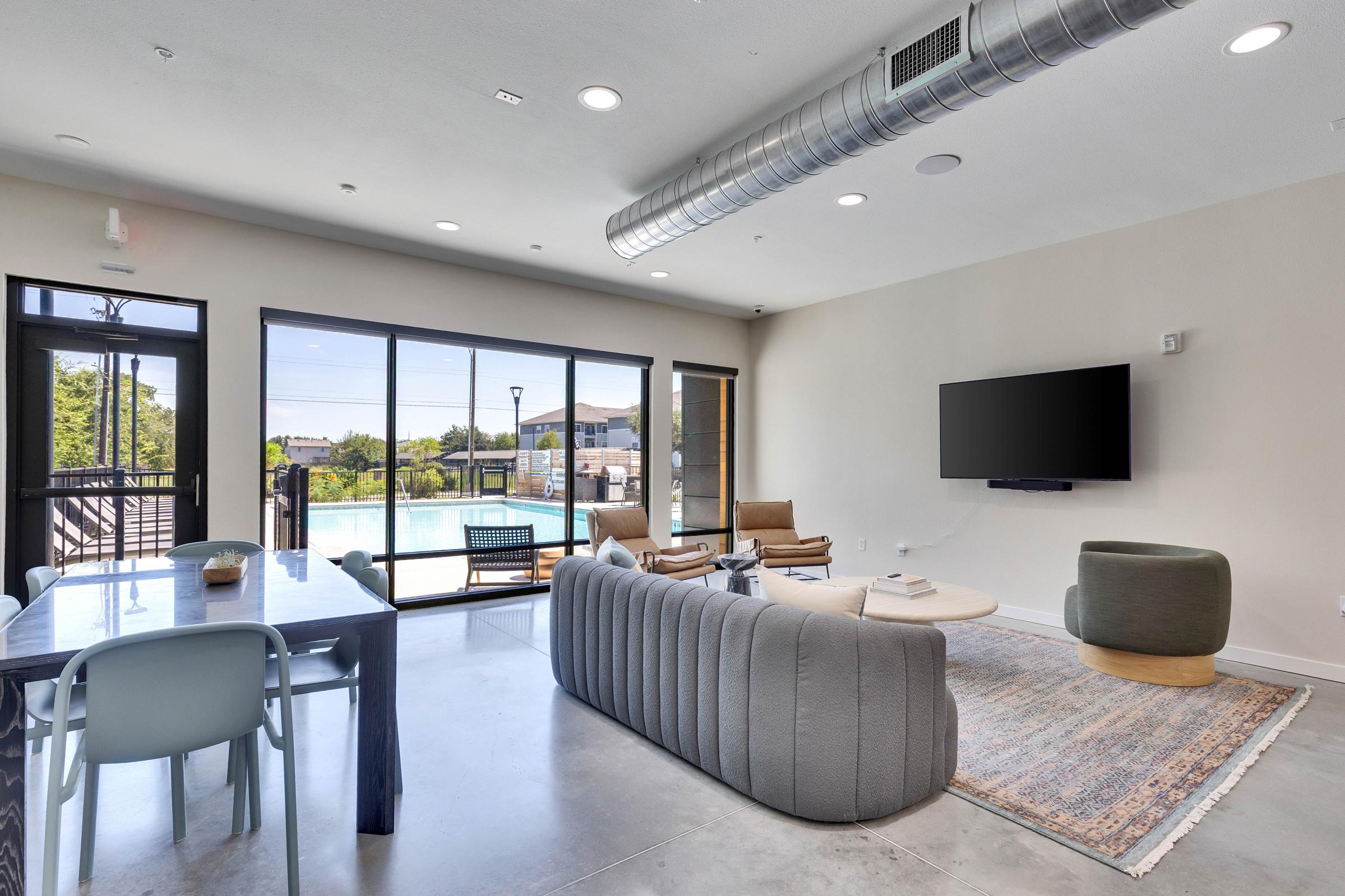 Modern interior of a spacious living area featuring large windows, a comfortable gray sofa, wooden armchairs, a round coffee table, and a dining table with chairs. A TV is mounted on the wall, and a pool area is visible outside, creating a bright and inviting atmosphere.
