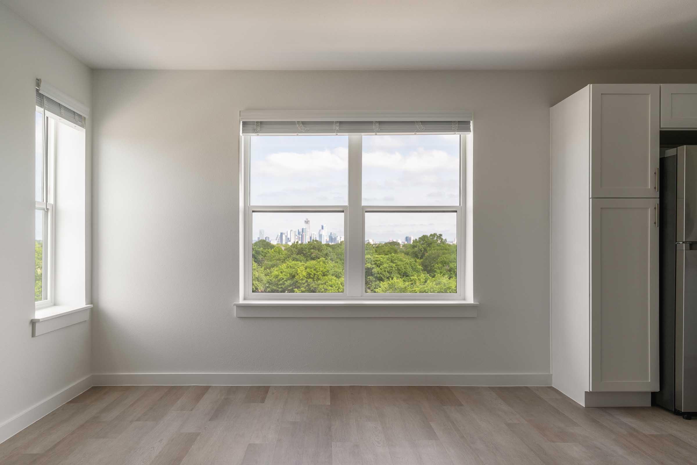 A bright and airy room with large windows offering a view of greenery and a city skyline. The walls are painted light gray, and the flooring is light wood. One corner of the room has white cabinetry, creating a modern and clean look. Natural light floods the space, enhancing its inviting atmosphere.