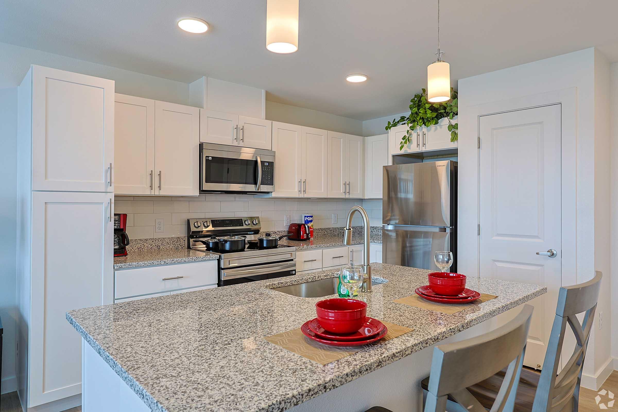 A modern kitchen featuring white cabinetry, stainless steel appliances, and a sleek granite countertop. The island has a sink and is set with two red plates and glasses. There are pendant lights overhead, and greenery decor adds a fresh touch. The space is bright and inviting, with a cozy dining setup.
