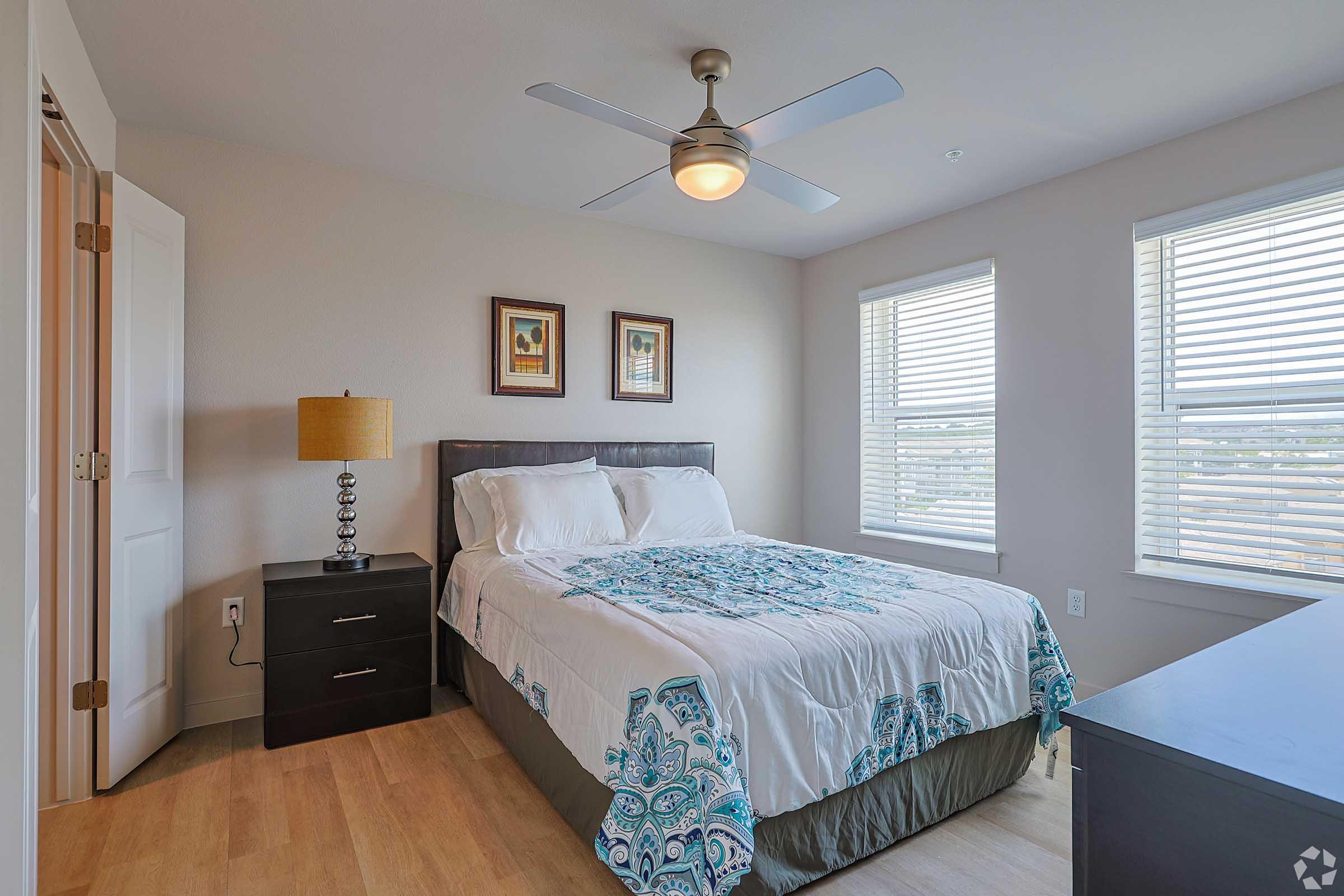 A cozy bedroom featuring a queen-sized bed with a decorative blue and white bedspread. There's a bedside table with a lamp, a modern ceiling fan, and two windows with blinds allowing natural light. Framed artwork adorns the wall above the bed, creating a welcoming atmosphere.