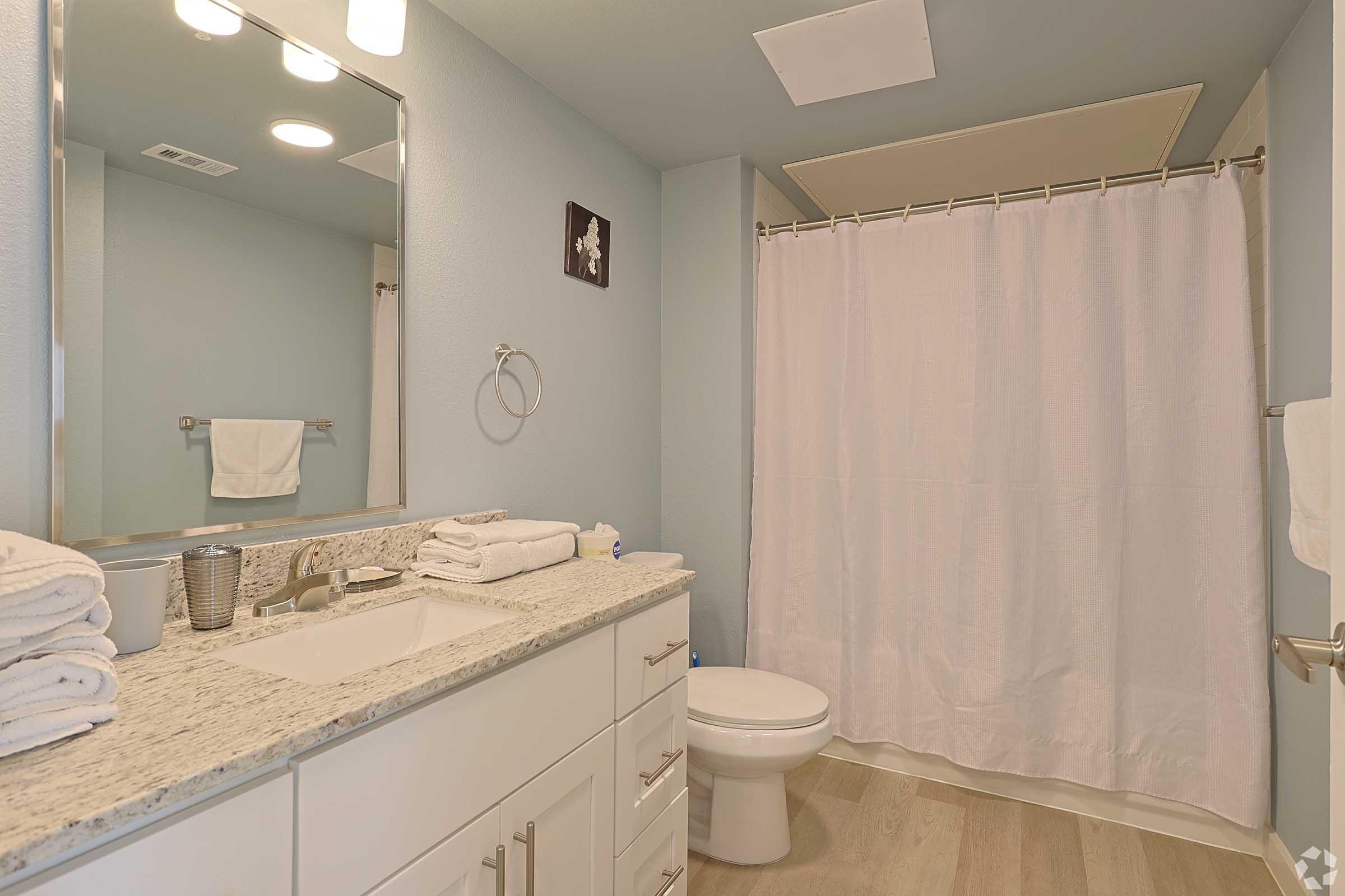 A clean, modern bathroom featuring a sink with granite countertop, neatly arranged towels, a white shower curtain, and a toilet. The walls are painted in a soft blue color, and there is a large mirror above the sink, with a well-lit ceiling fixture providing illumination.