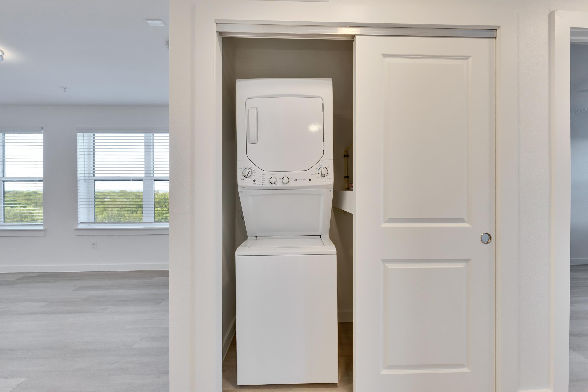 A compact laundry unit featuring a stacked washer and dryer is tucked into a closet space with double doors. The room has large windows allowing natural light, and the floor has a light wood finish, suggesting a modern and open living area.