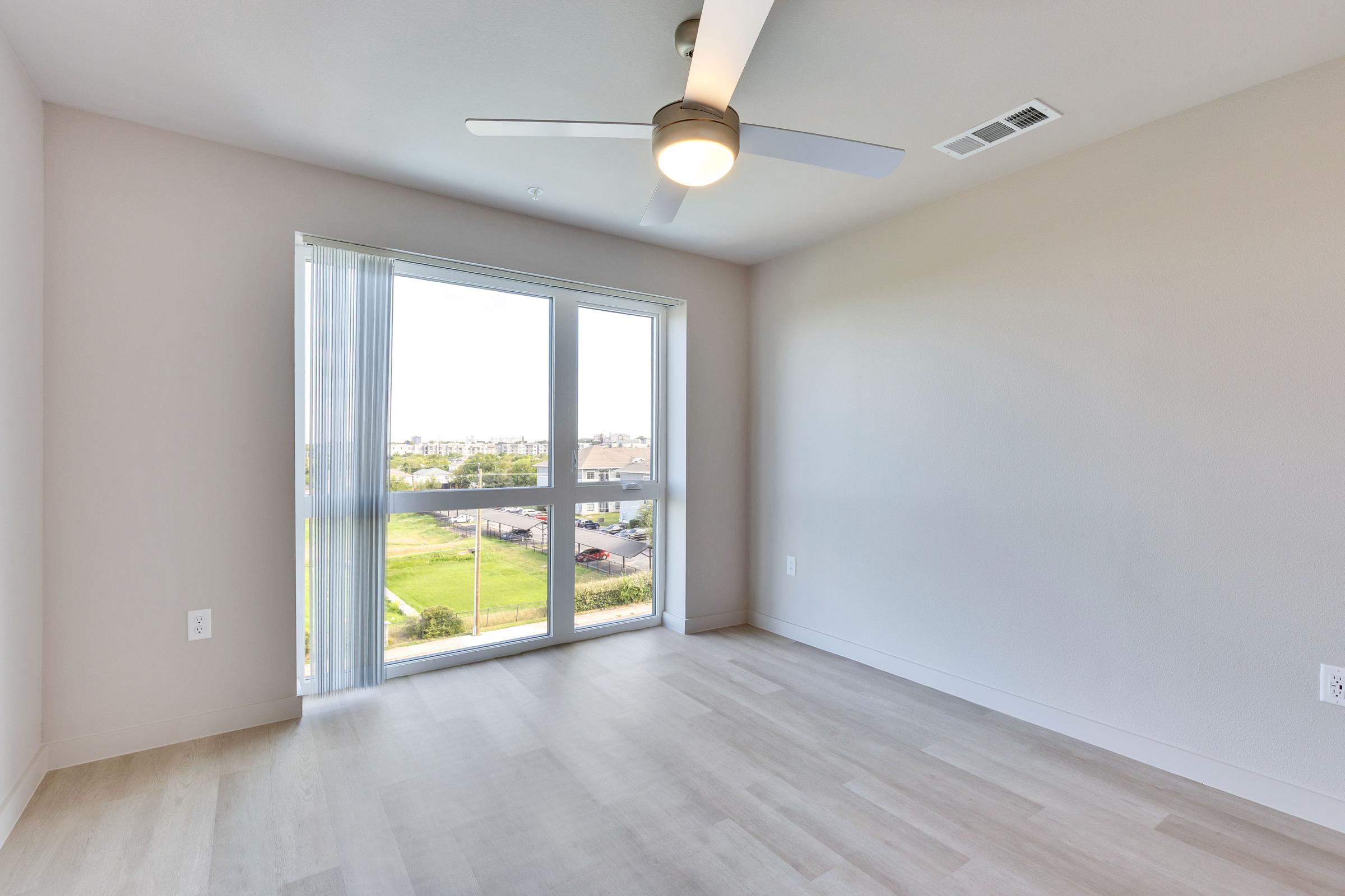Interior of a light and airy empty room featuring large windows with sheer curtains, a ceiling fan, and wood-like flooring. The view outside shows greenery and residential buildings. The walls are painted in a soft neutral color, creating a bright and inviting atmosphere.