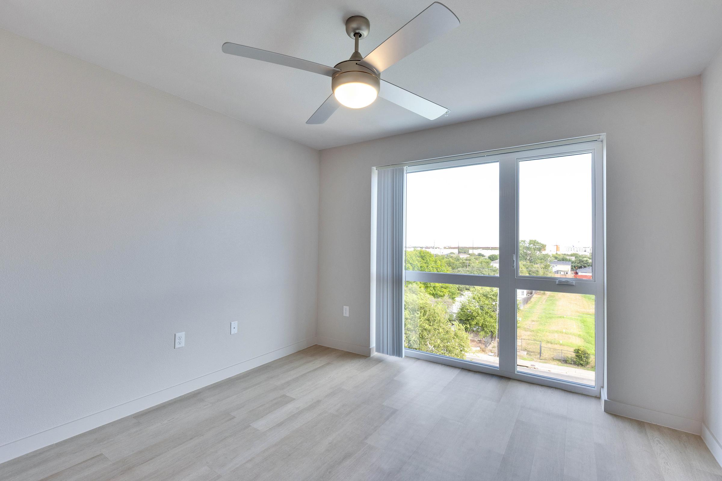 Empty room with light hardwood flooring and a large window providing natural light. A modern ceiling fan is visible, and the walls are painted in a neutral color. The view outside includes greenery and a glimpse of the skyline.