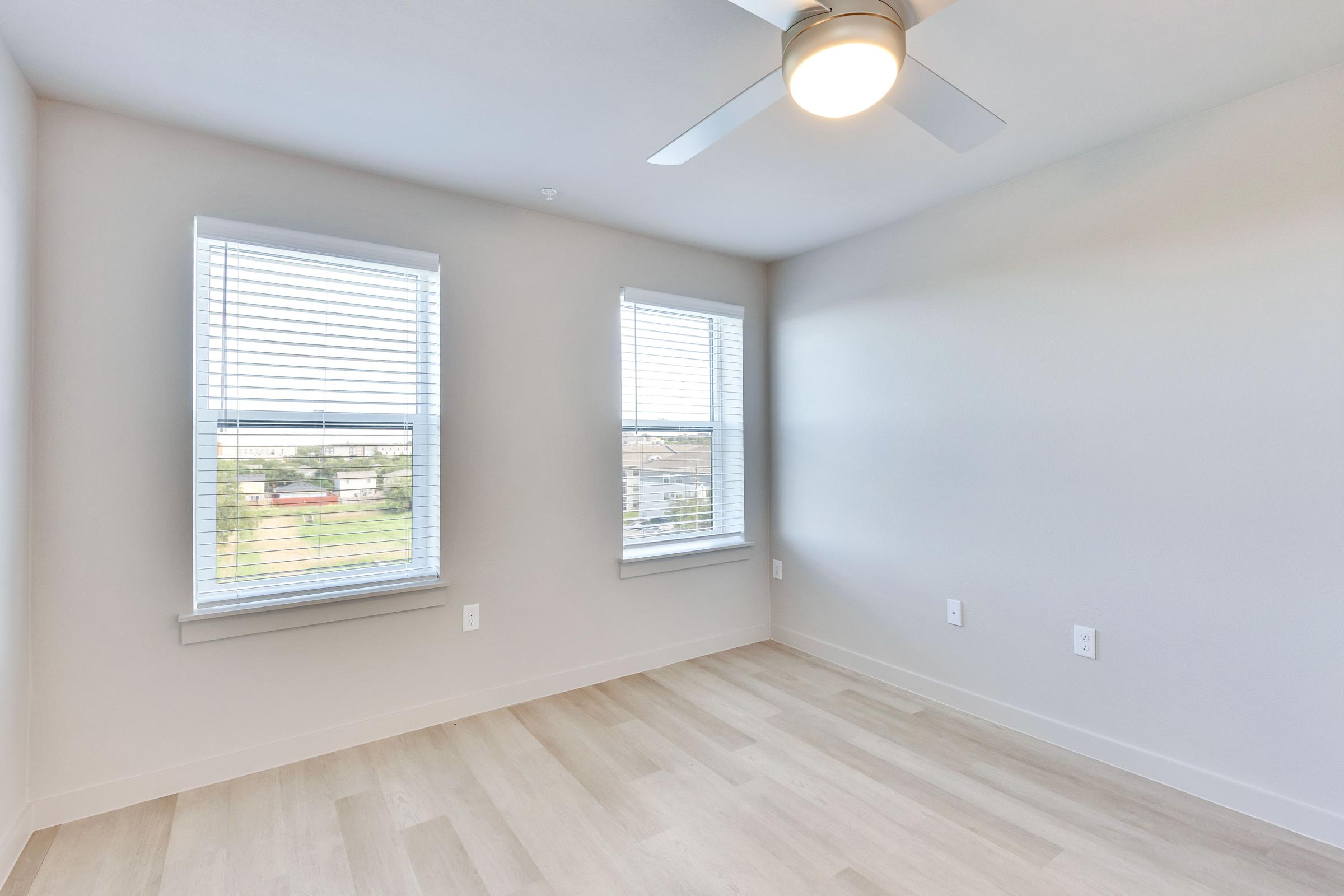 A bright, empty room with light-colored walls and wooden flooring. Two windows with white blinds allow natural light to enter, and a ceiling fan is mounted in the center. The view outside appears to be a grassy area, creating a serene atmosphere.