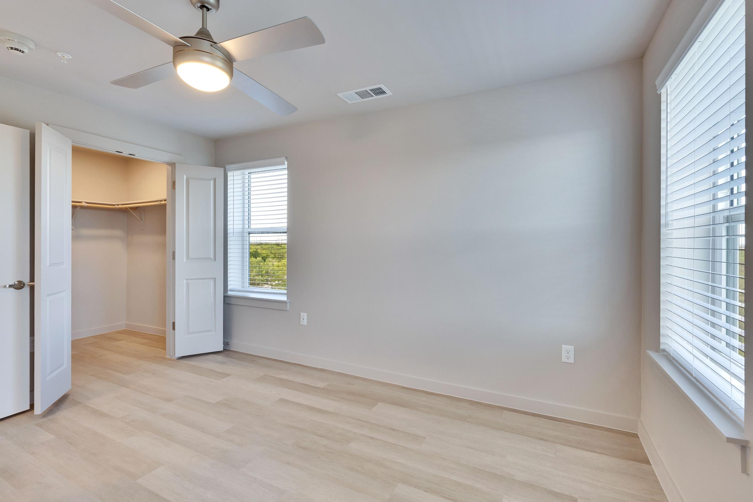 A bright, empty bedroom featuring light-colored walls and flooring, a ceiling fan, and large windows with blinds. One closet door is open, revealing an empty closet space. The room is well-lit and has a modern, minimalist design.