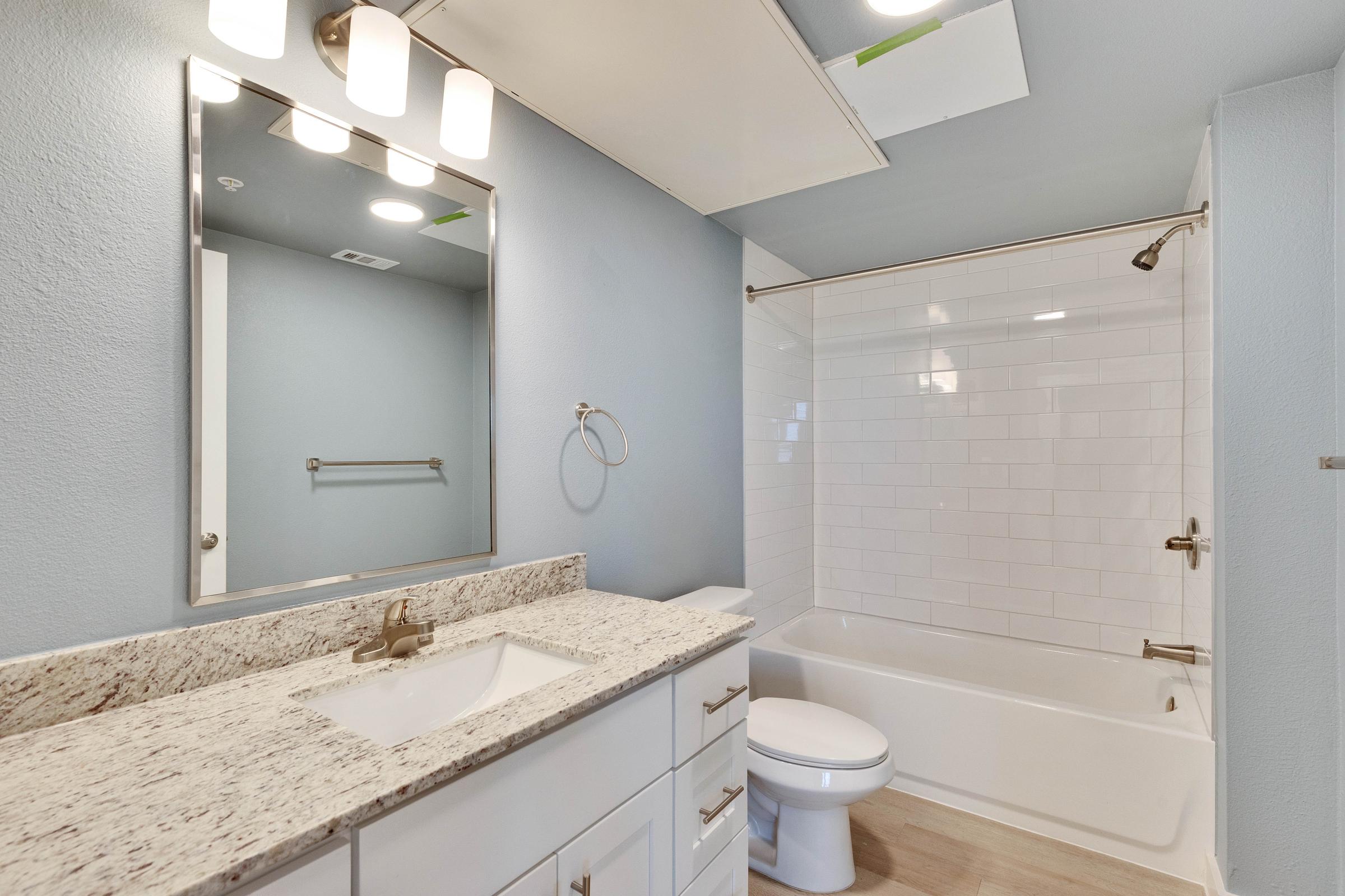 A modern bathroom featuring a light blue wall, white subway tile bathtub, and a white vanity with a granite countertop. The space includes a rectangular mirror above the sink, a towel rack, and bright overhead lighting. The overall design is clean and contemporary.