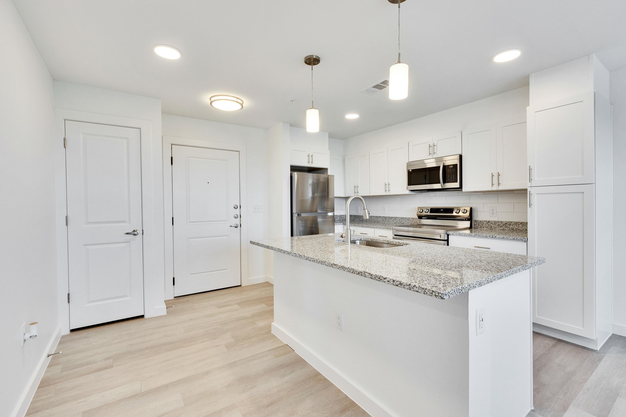 A modern kitchen featuring white cabinets, stainless steel appliances, and a large island with a granite countertop. The open layout includes light wood flooring and bright overhead lighting. A door leads to the entrance, enhancing the spacious feel of the area.
