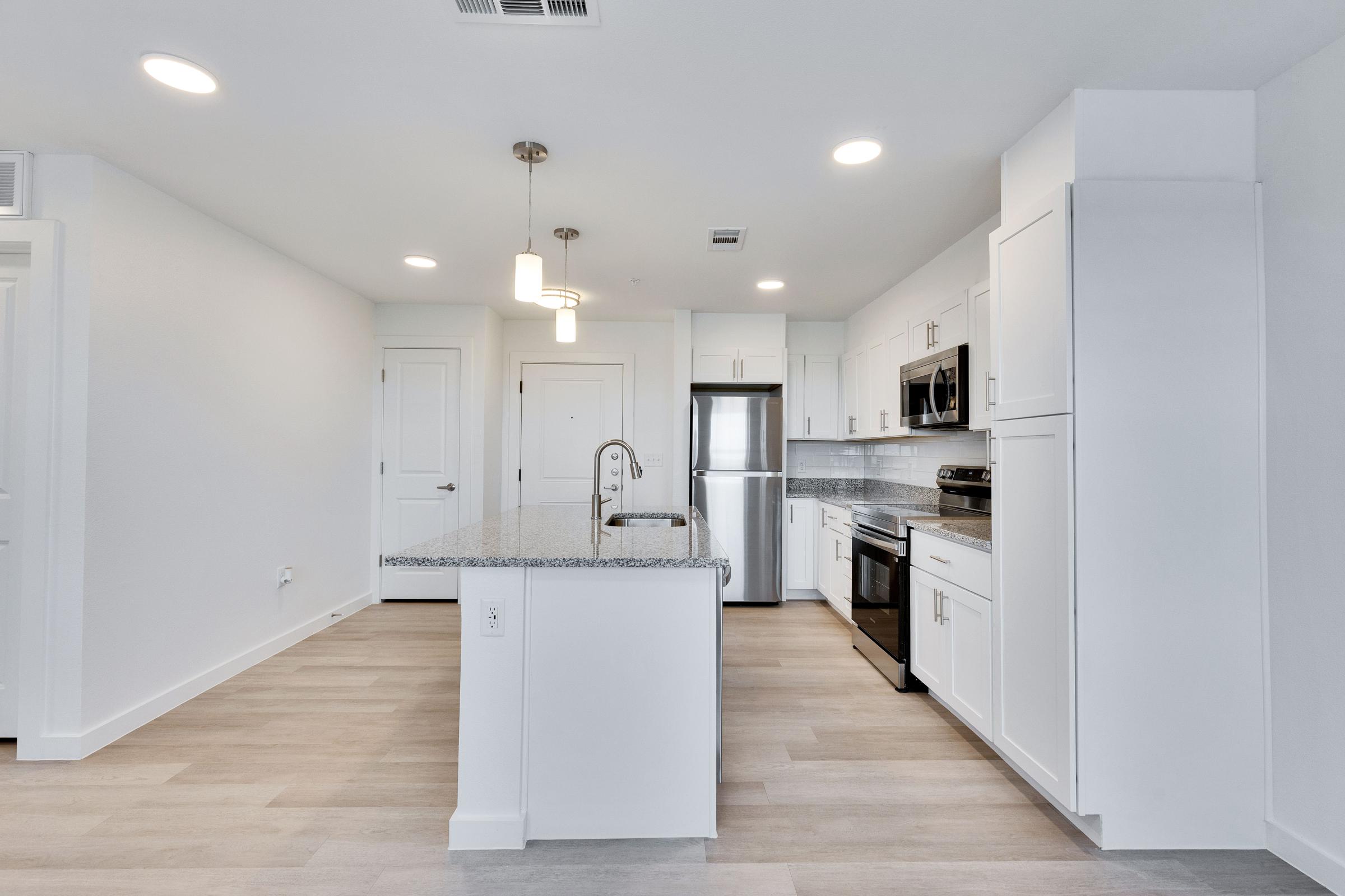 Modern kitchen with white cabinetry, stainless steel appliances including a refrigerator and microwave, and a large island with a gray countertop. The space features light-colored wooden flooring and bright overhead lighting, creating a clean and inviting atmosphere.