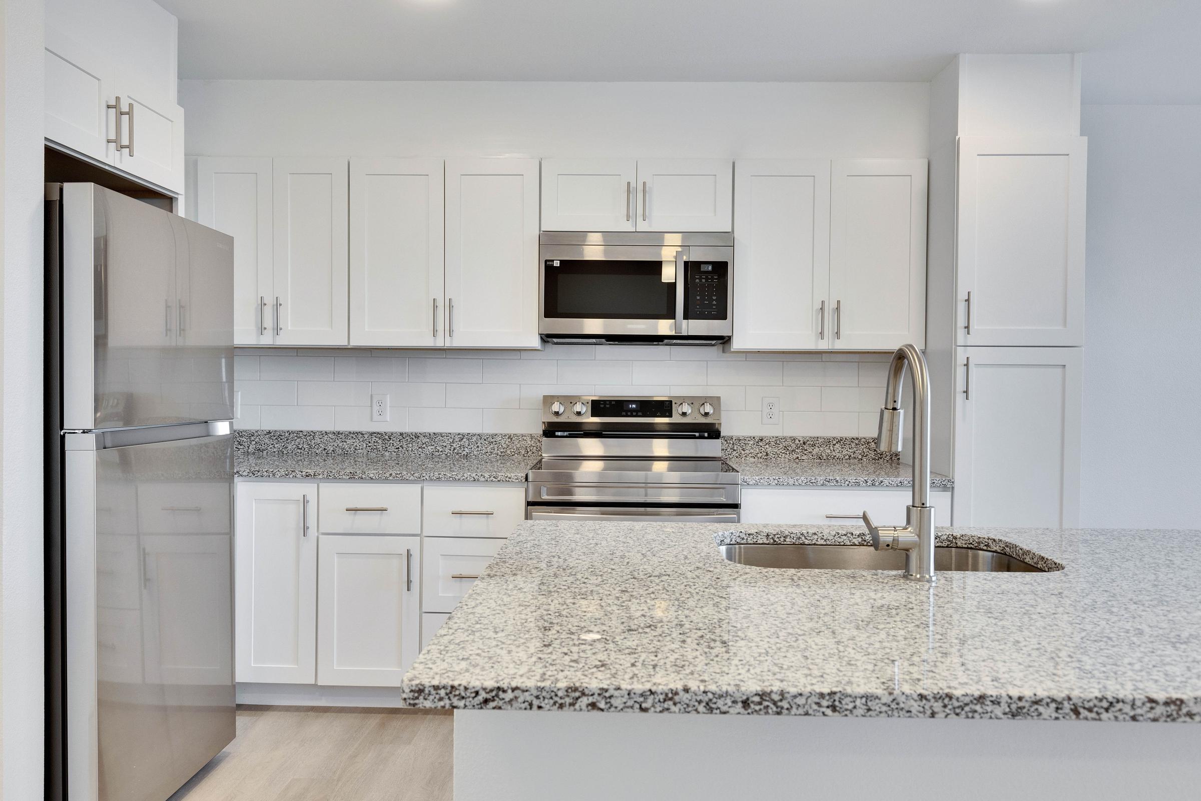 Modern kitchen featuring white cabinets, a stainless steel refrigerator, an oven with a microwave above, and a large granite countertop with an undermount sink. The space is bright and sleek, showcasing a clean and contemporary design with light-colored flooring and minimalistic fixtures.